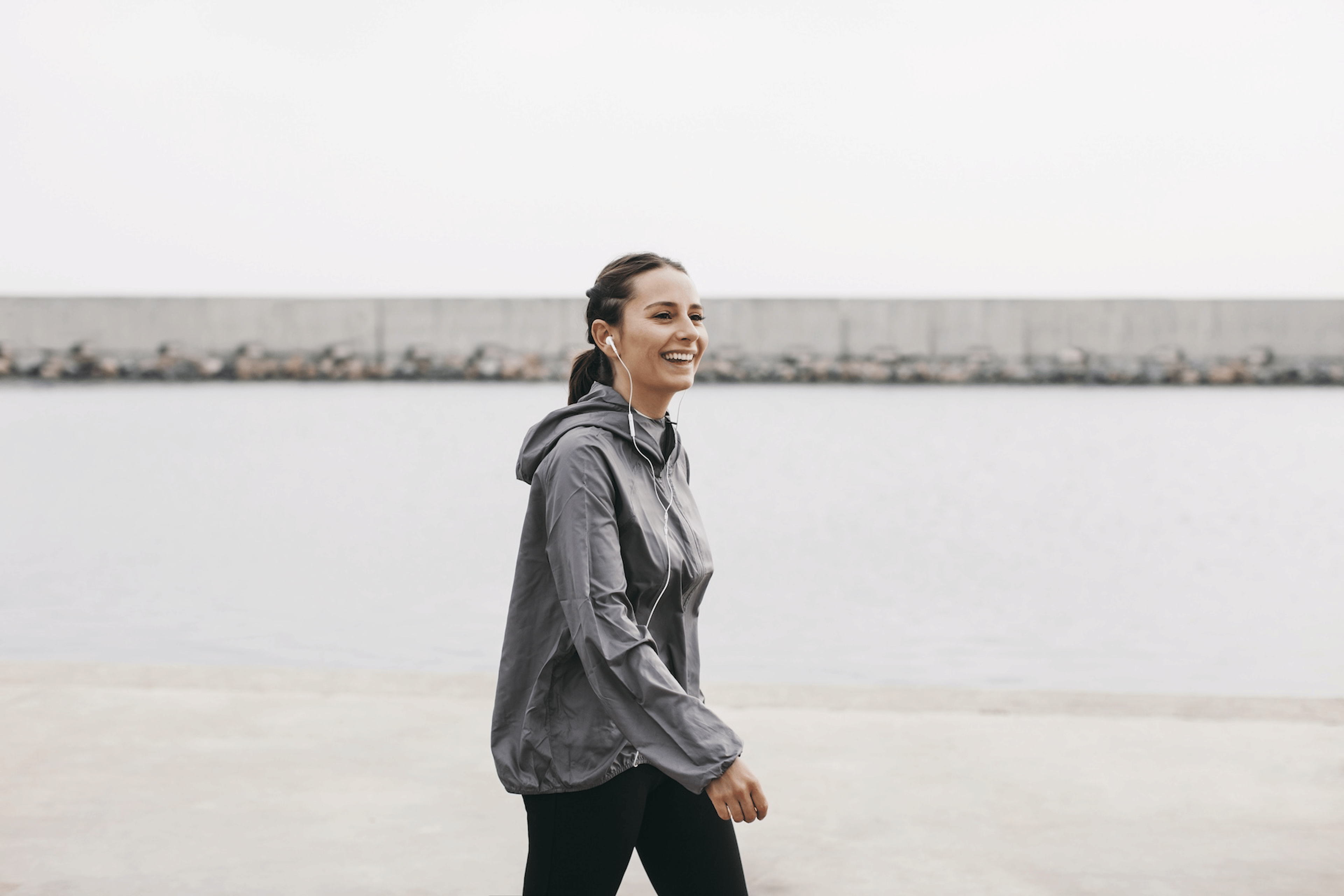 A happy woman walking outside near a harbor. She has headphones in.