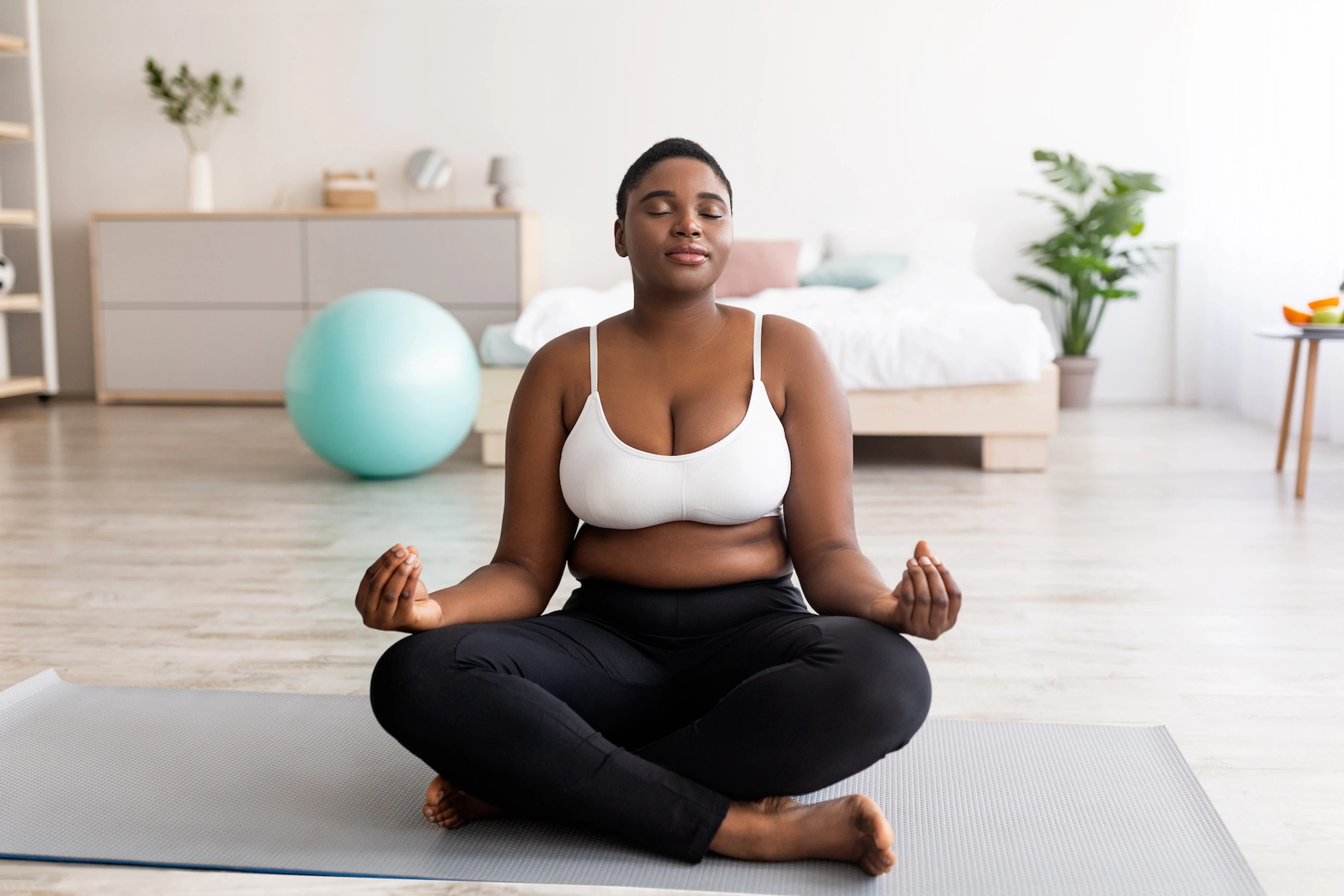 A woman sitting on a yoga mat at home doing a postpartum workout. She is in lotus position with her eyes closed.
