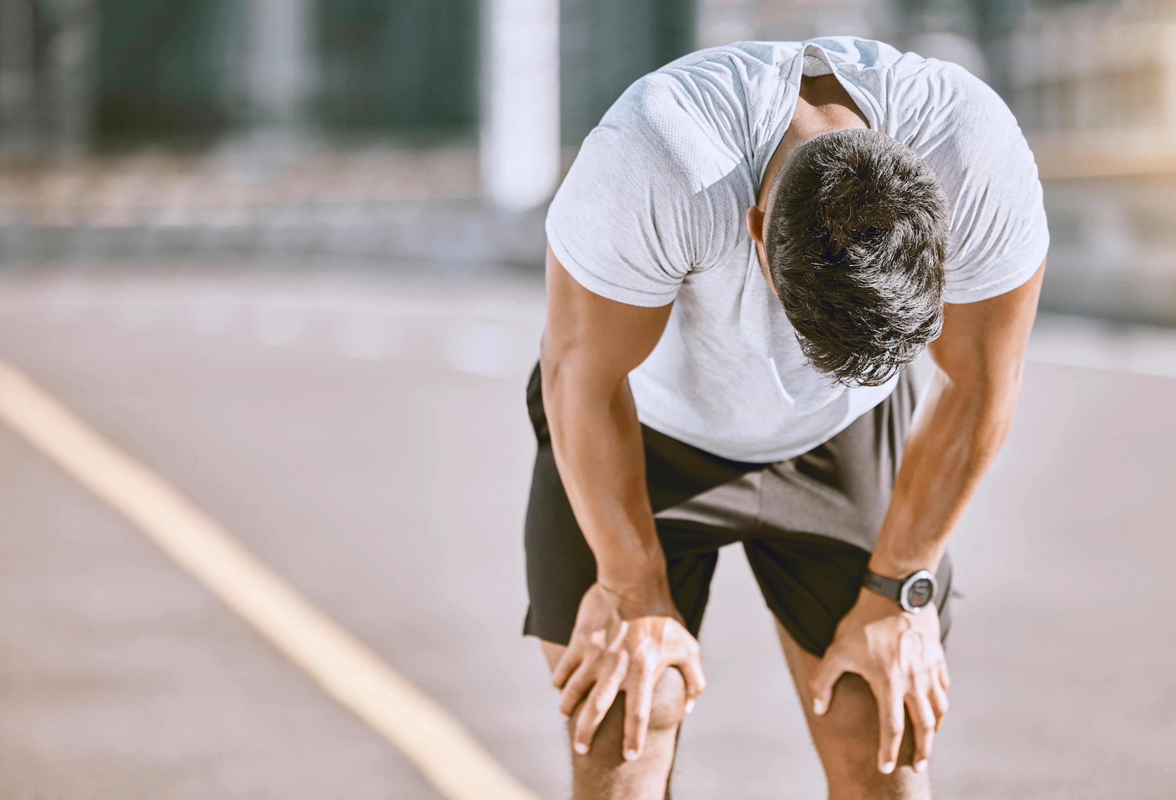 A man feeling lightheaded after a workout. His hands are on his knees and he is tilting his head down after an outdoor run.