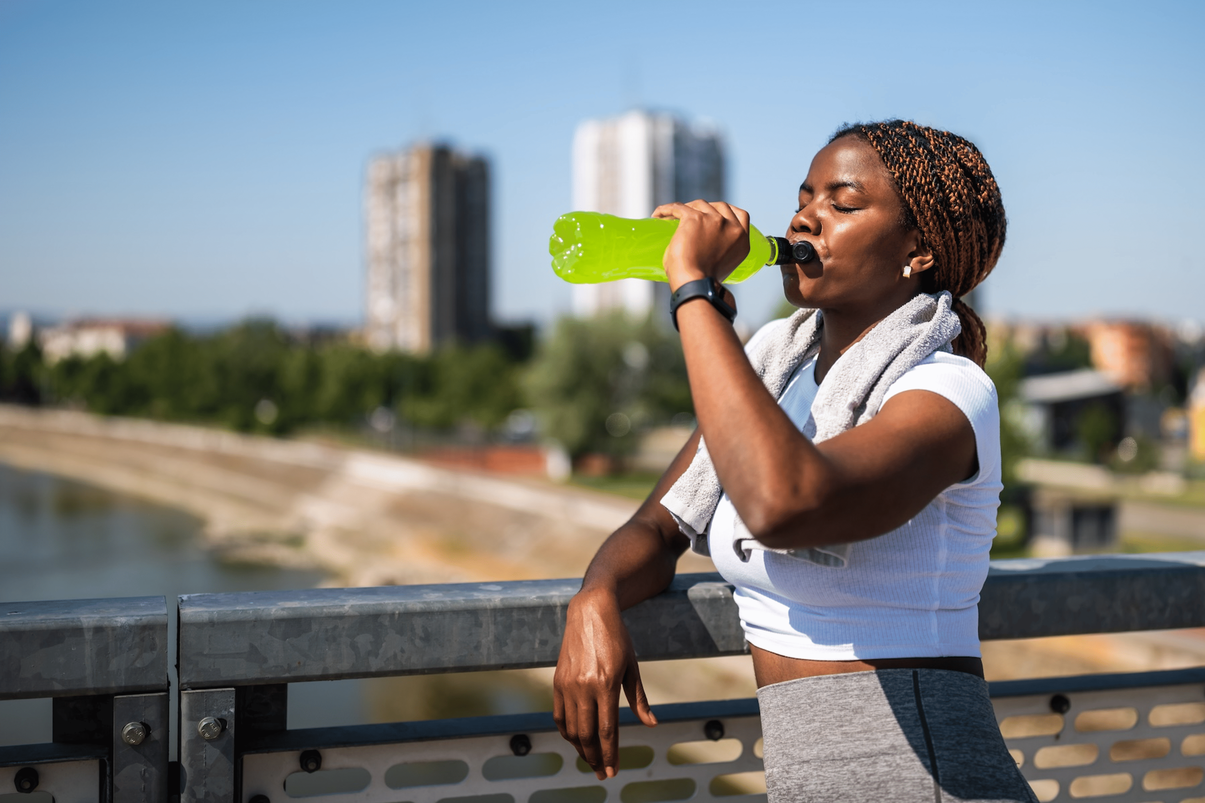 A runner leaning against a fence and drinking a green sports drink to replenish electrolytes on during a hot, sunny run.