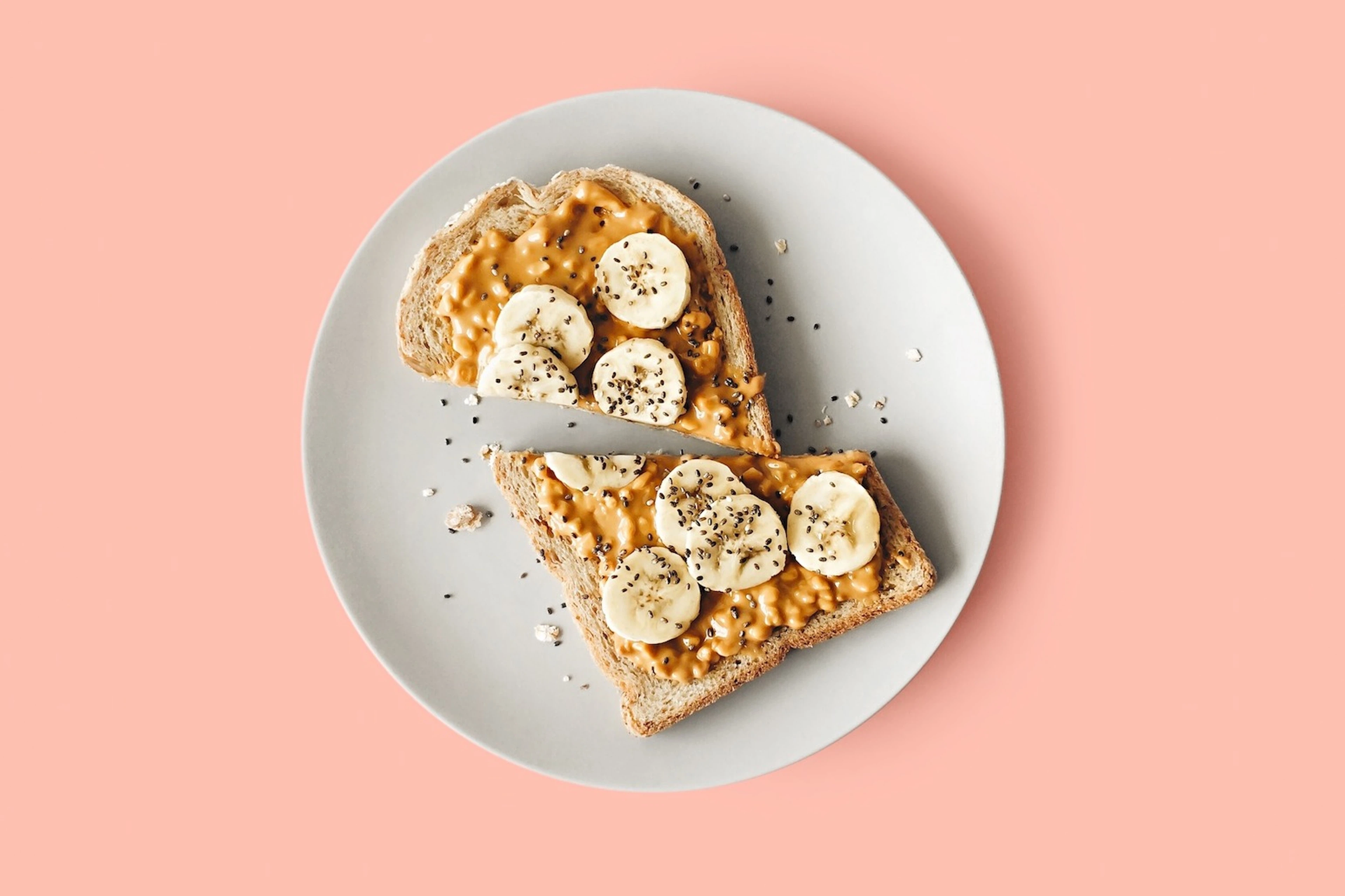 Two slices of peanut butter banana toast with chia seeds on a white plate resting on a pink background.