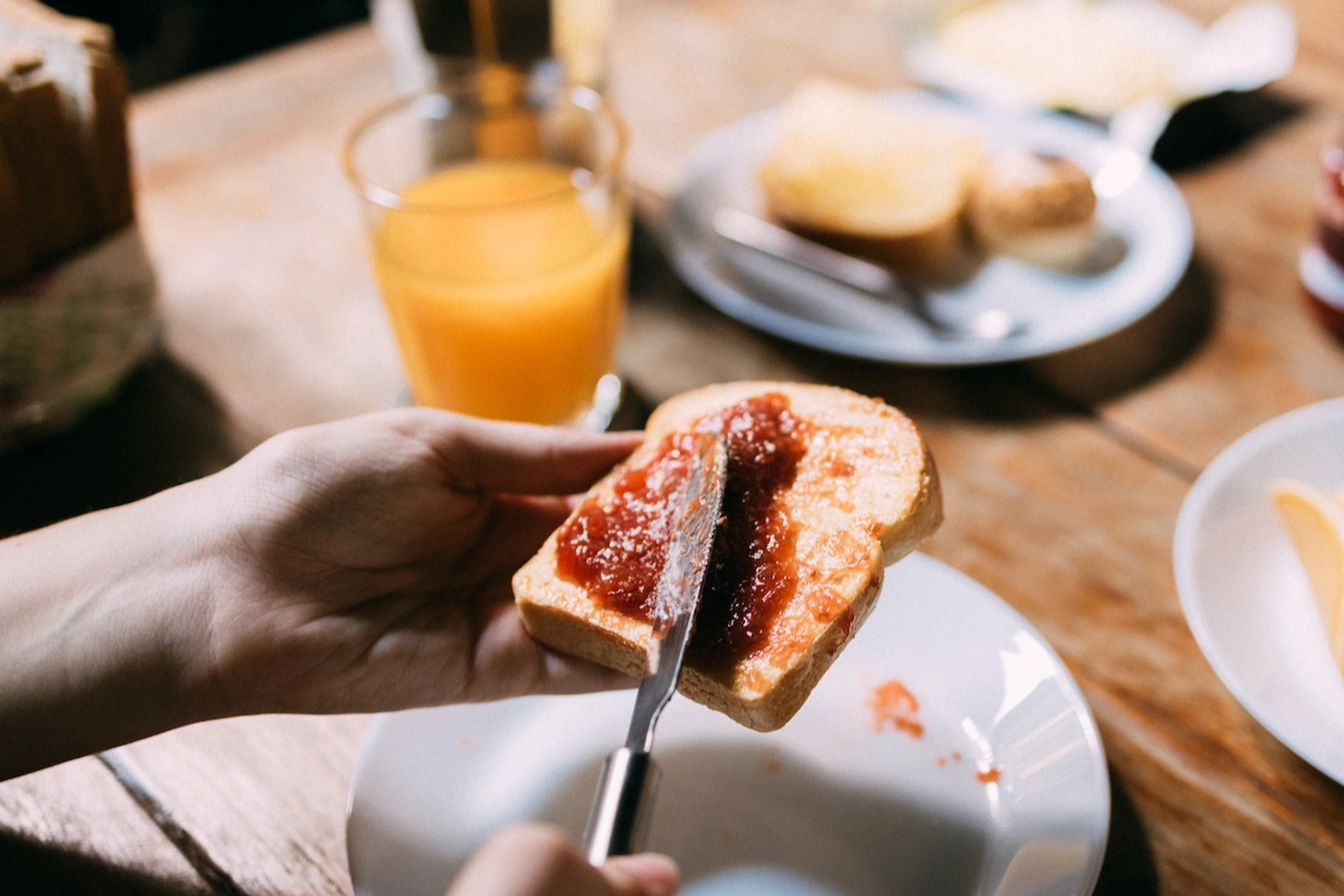 A person spreading jam on a piece of toast next to a glass of orange juice. This meal would make an ideal breakfast before a marathon.