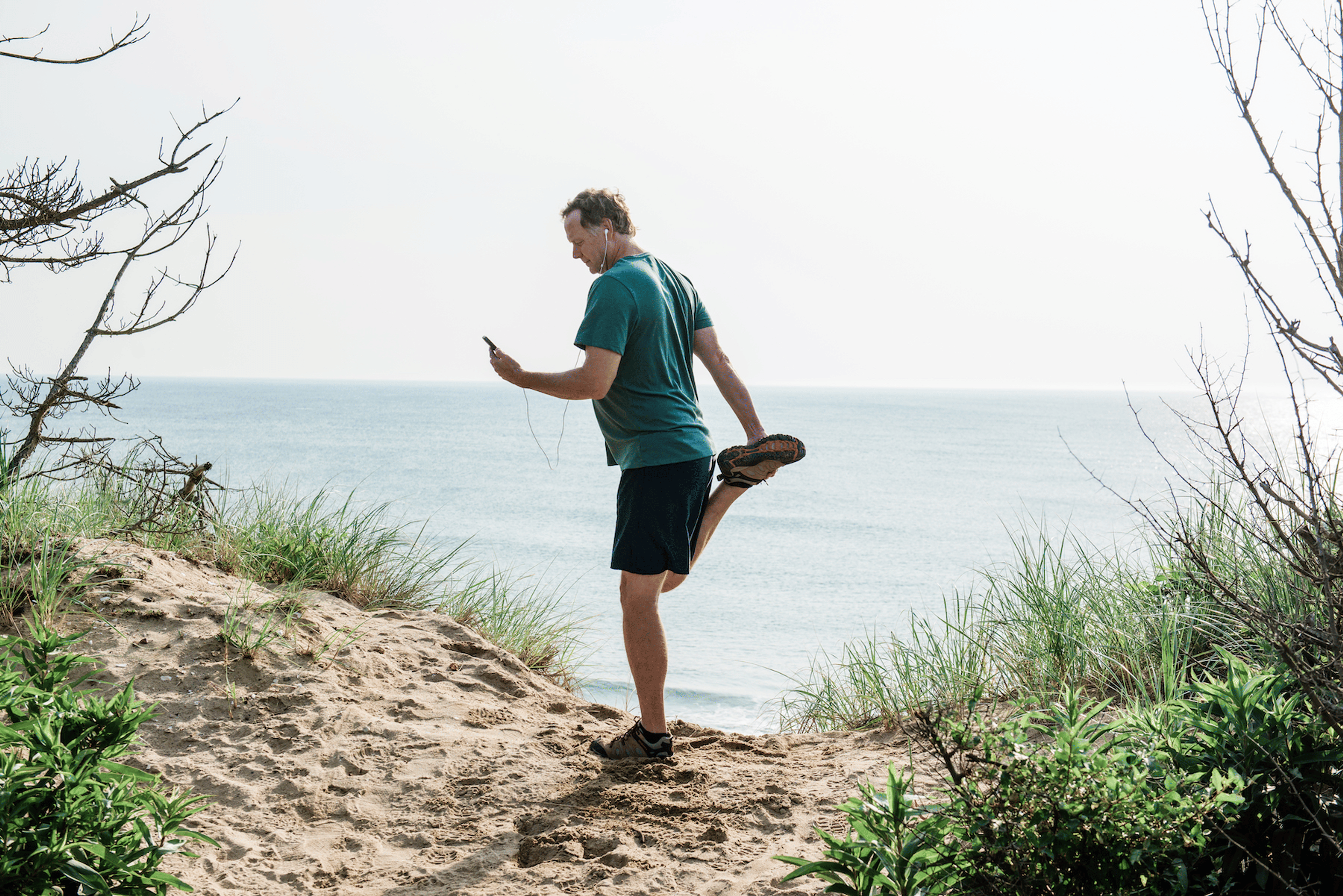 A man watching a video on his phone with wire headphones while simultaneously doing a standing quad stretch. He is standing on a beautiful sandy trail near the ocean.