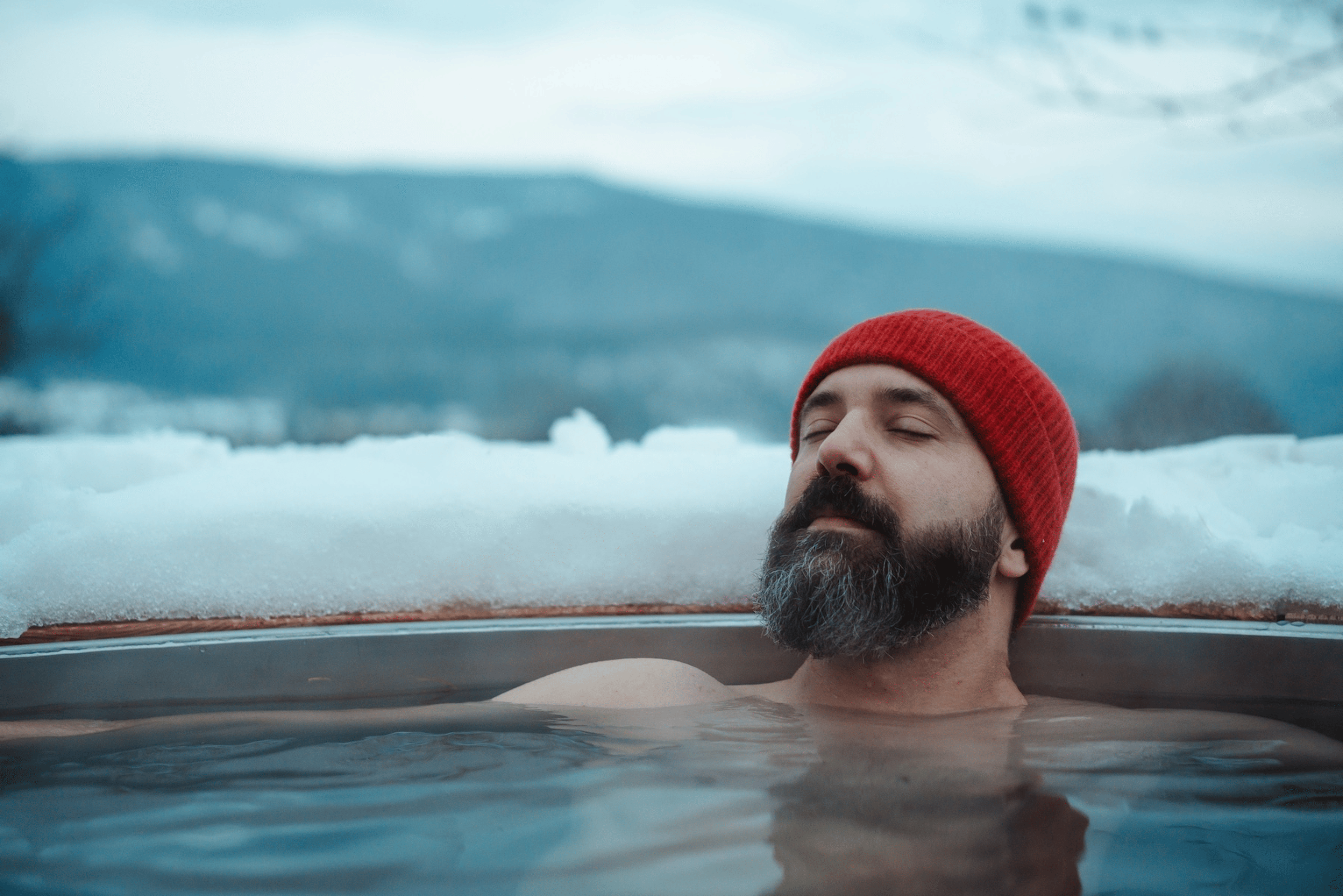 A man resting in a hot tub in a snowy outdoor environment. He's wearing a red beanie.