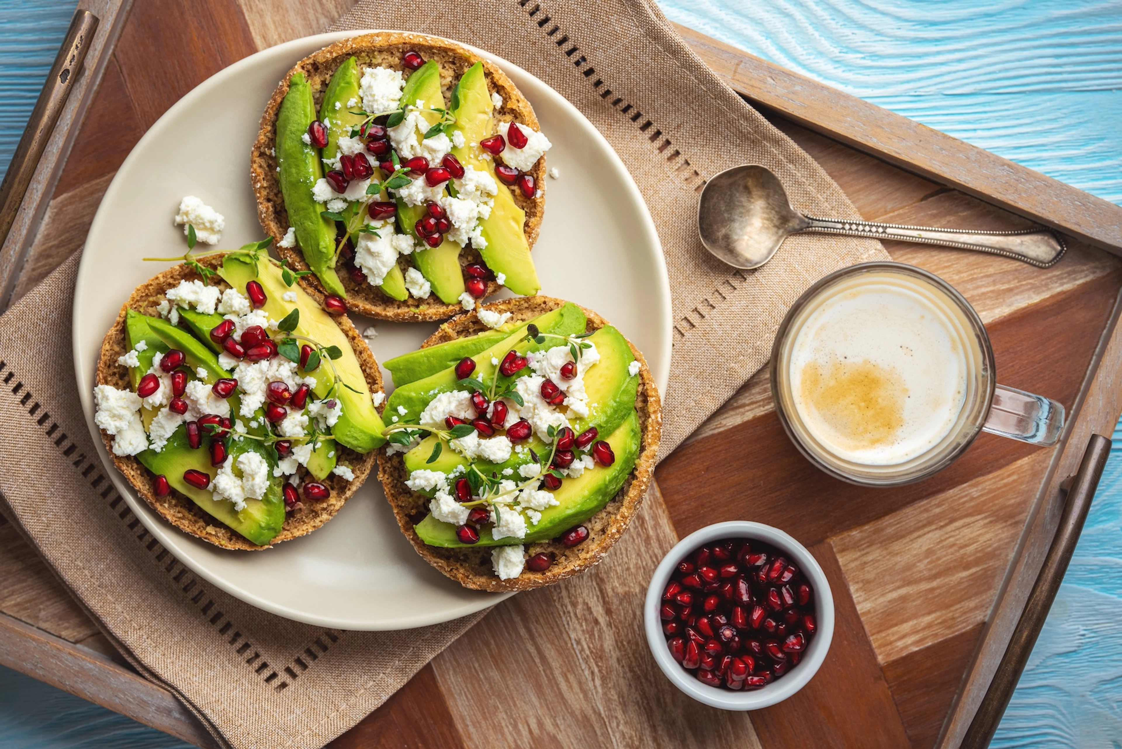 A plate of flat circular pieces of bread with avocado, pomegranate seeds, and feta cheese with a latte and extra bowl of pomegranate arils on the side.