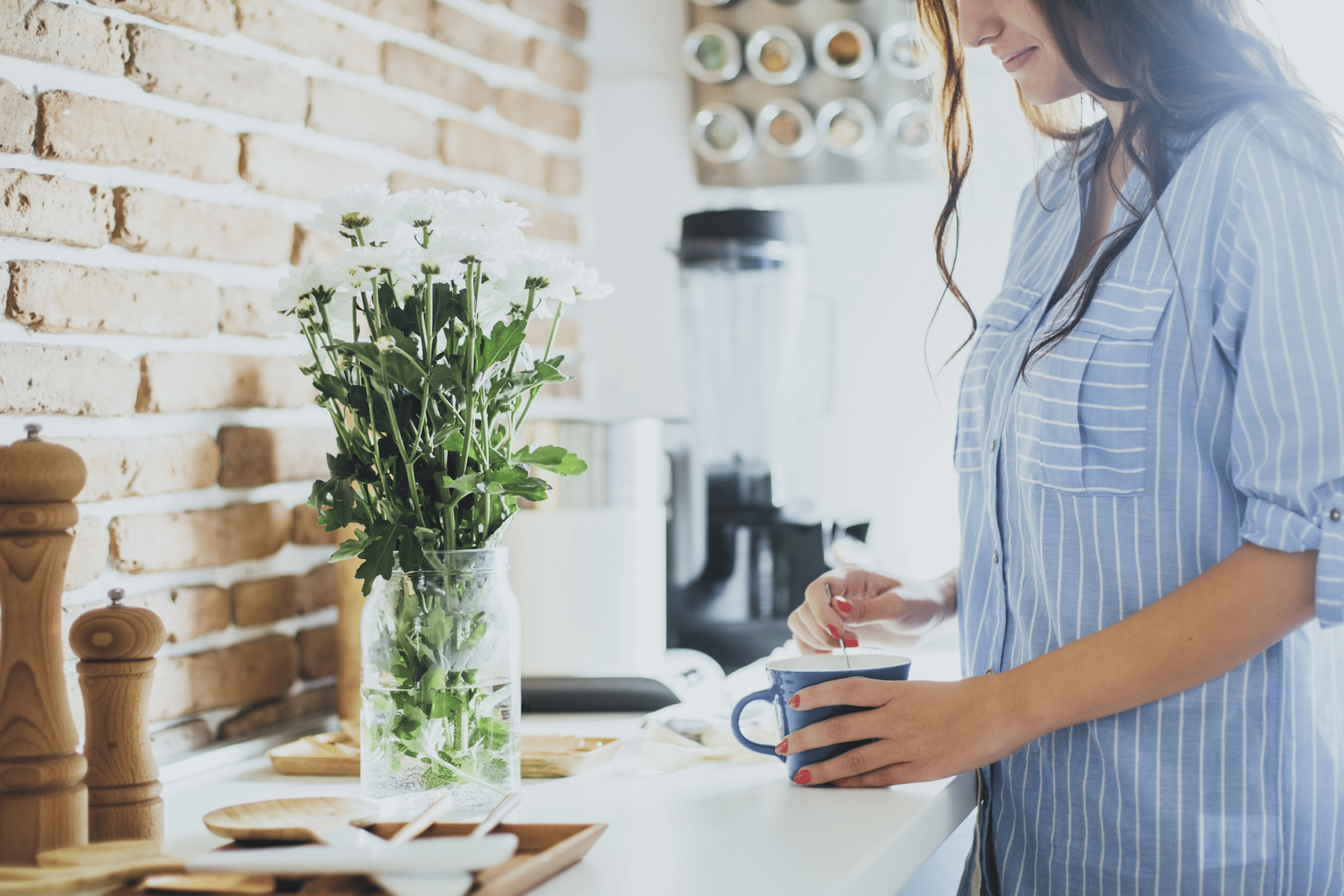 A woman stirring a cup of coffee in her sunny kitchen. There's a vase of fresh herbs next to her on the counter.