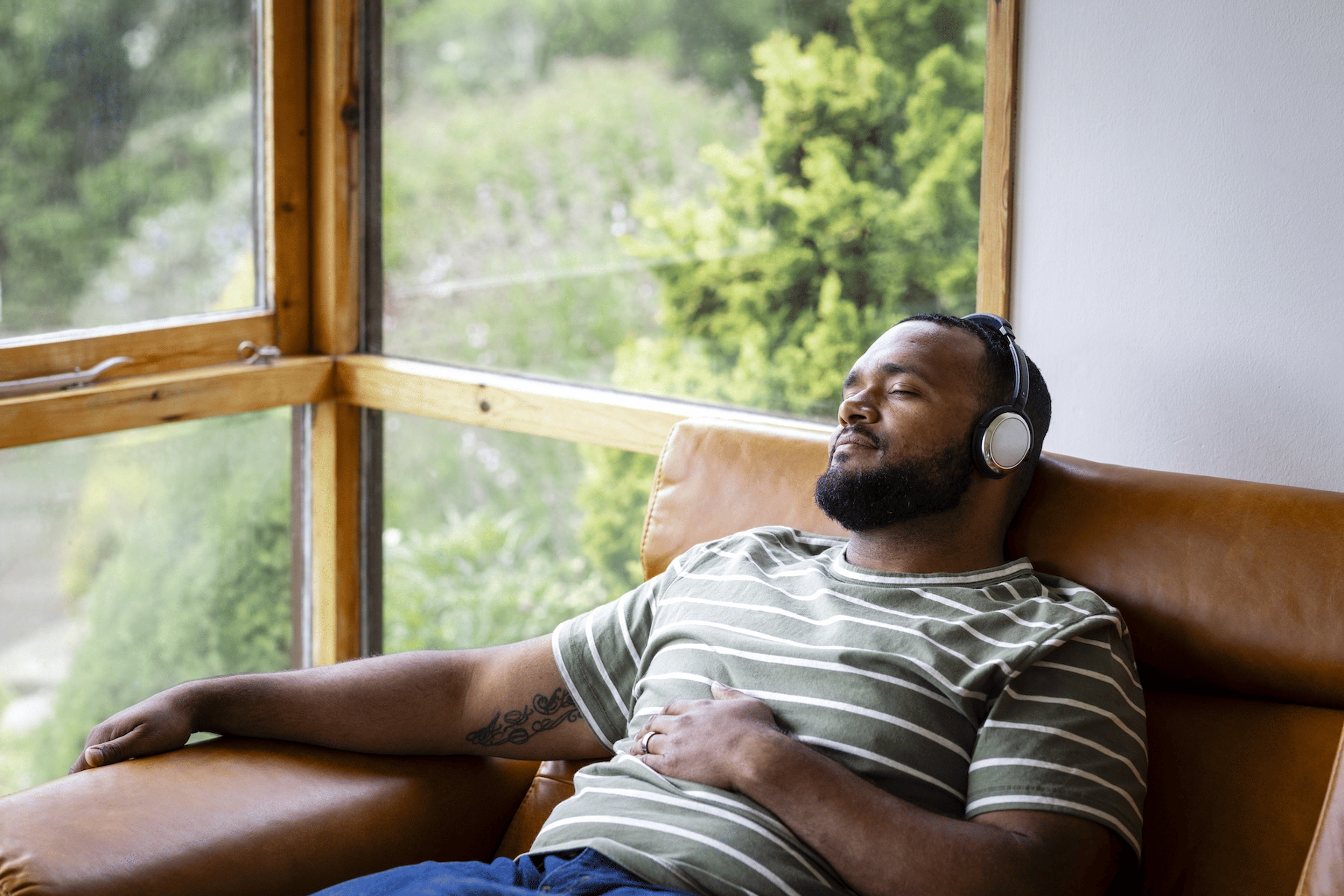 A man relaxing on a chair and listening to a guided meditation on his headphones. His eyes are closed and his left arm is resting on his belly.