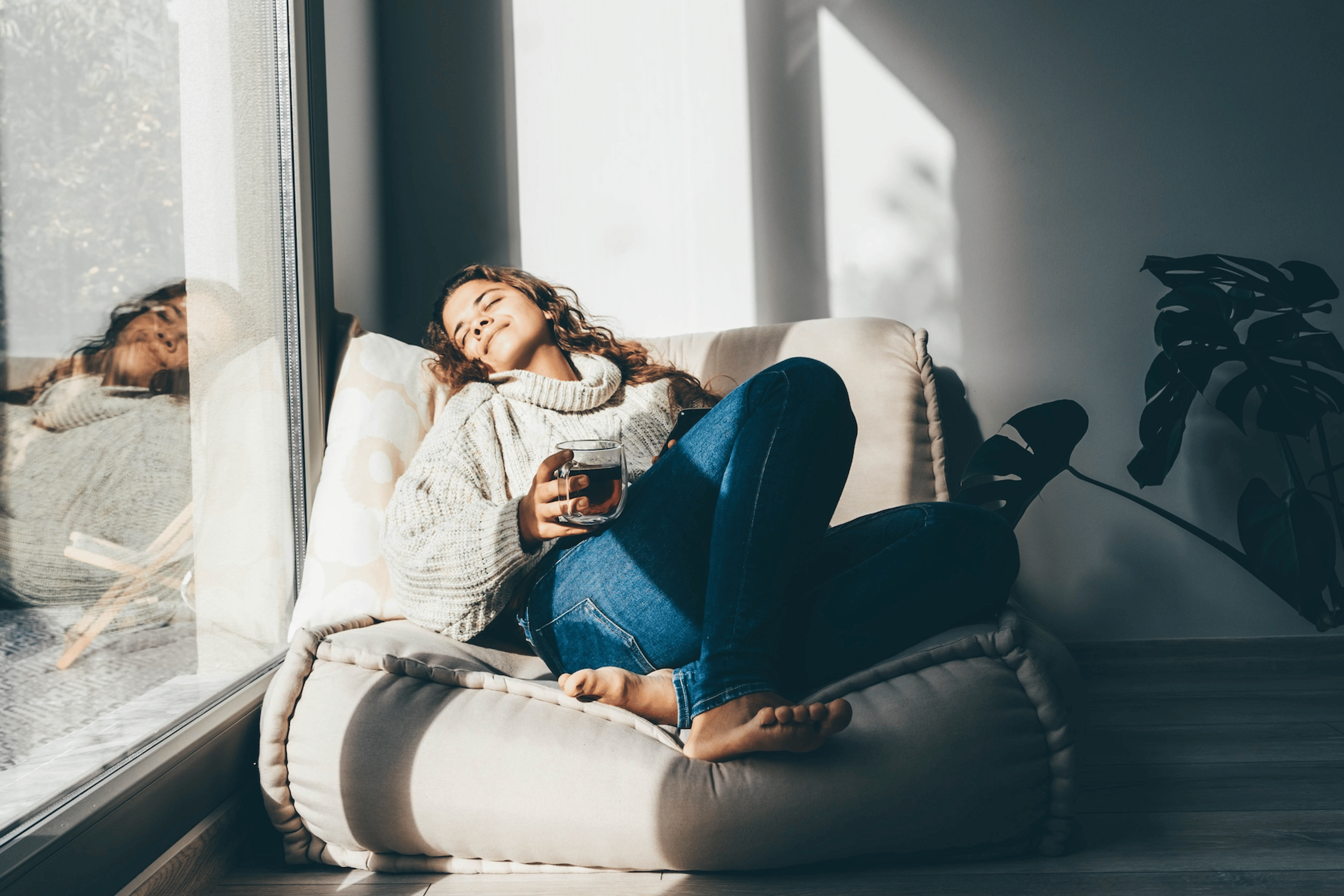A woman practicing self-care. She's resting on a comfy chair, basking in the sun, and drinking tea.