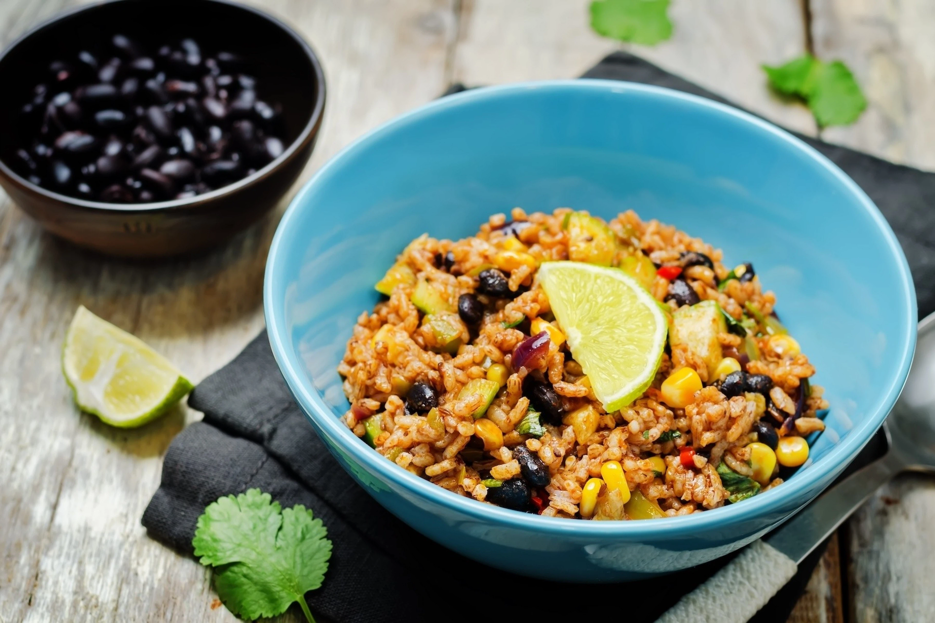 A bowl with black beans (a non-meat protein source) with zucchini and brown rice. A lime is garnishing the bowl and there is a bowl of black beans in the background out of focus.