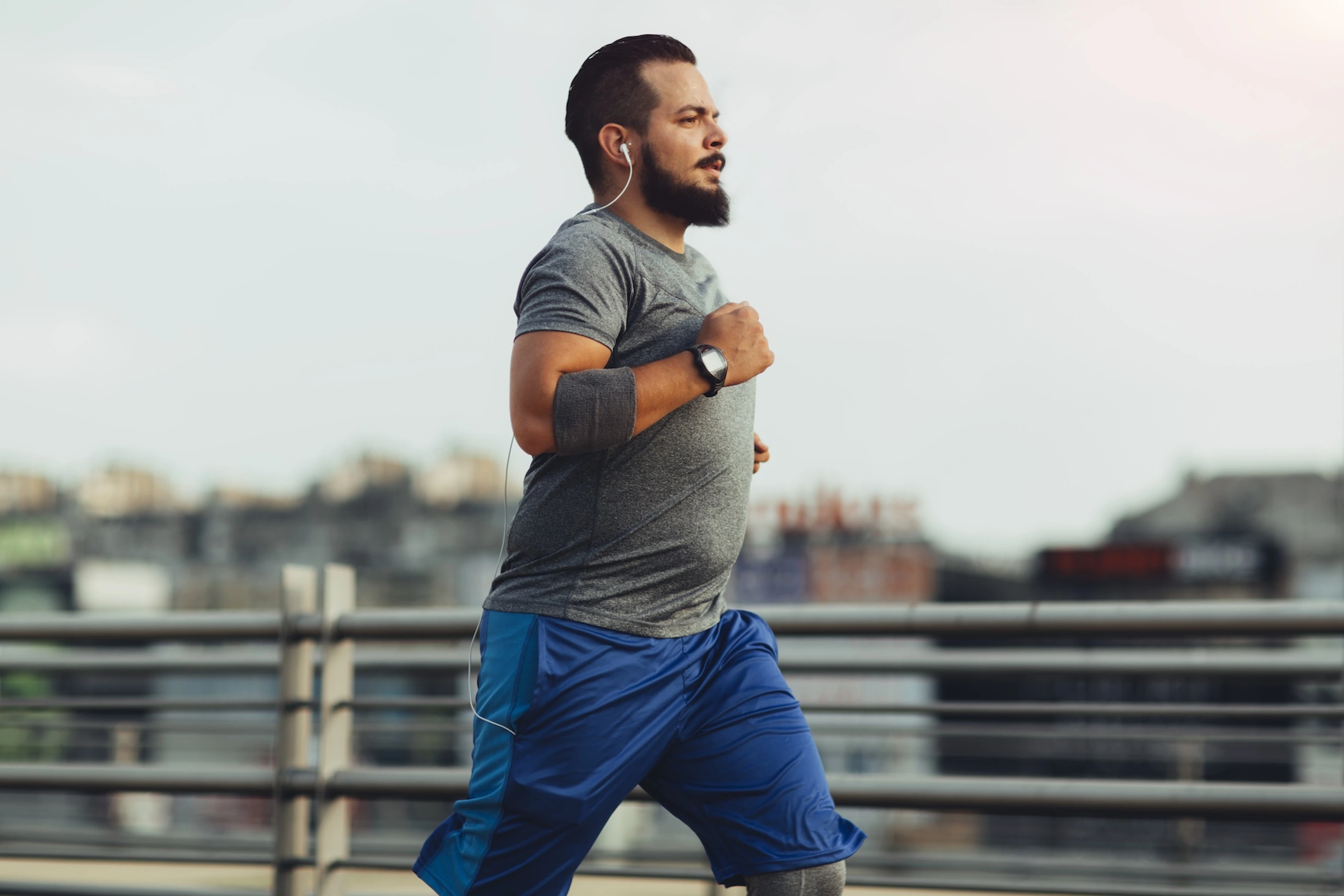 A man running outside to help ease anxiety. He's listening to his headphones and is wearing a gray tee, blue shorts, and a gray arm band.