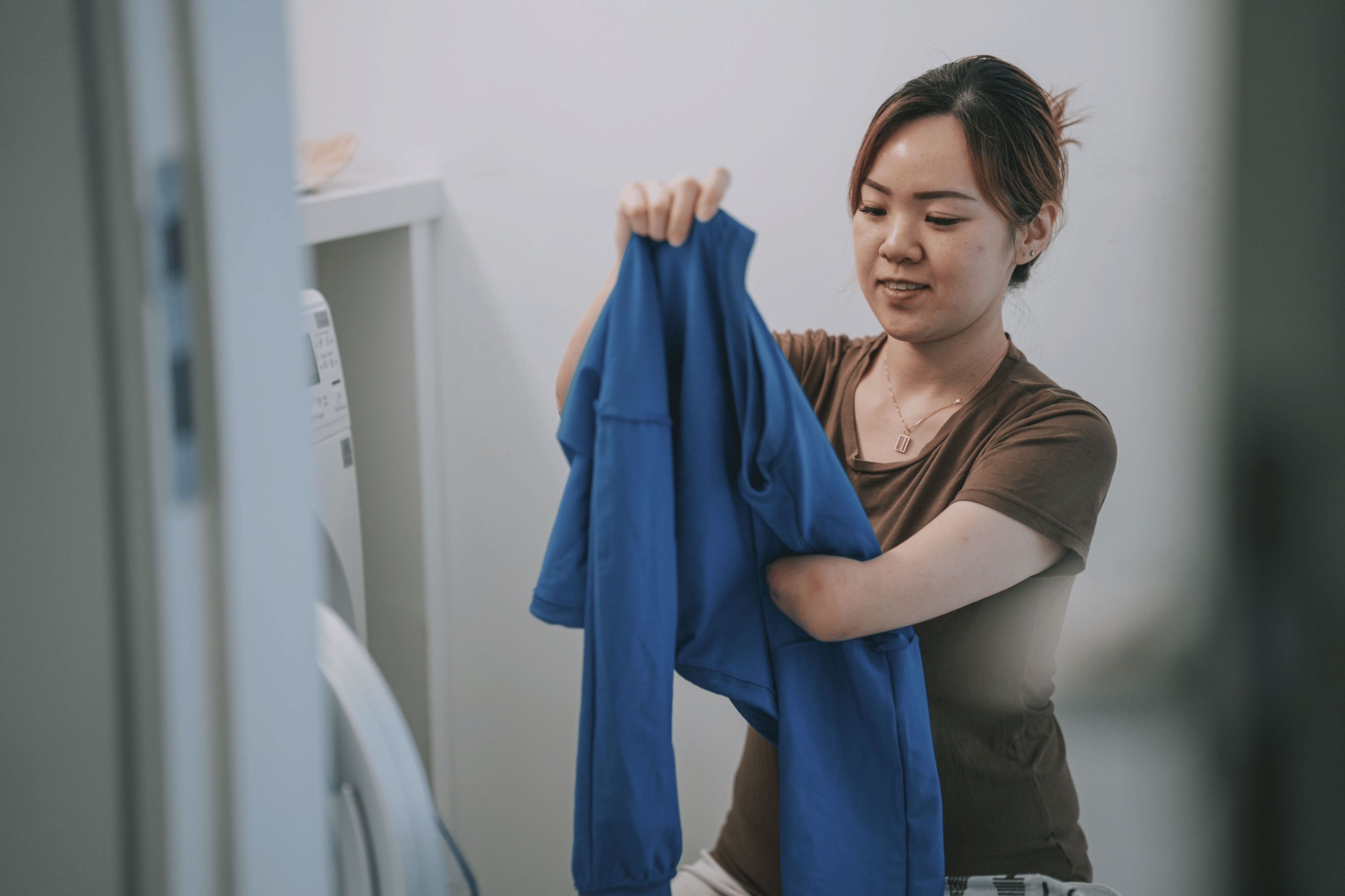 A woman holding up and folding a blue long-sleeve shirt at home in the laundry room.