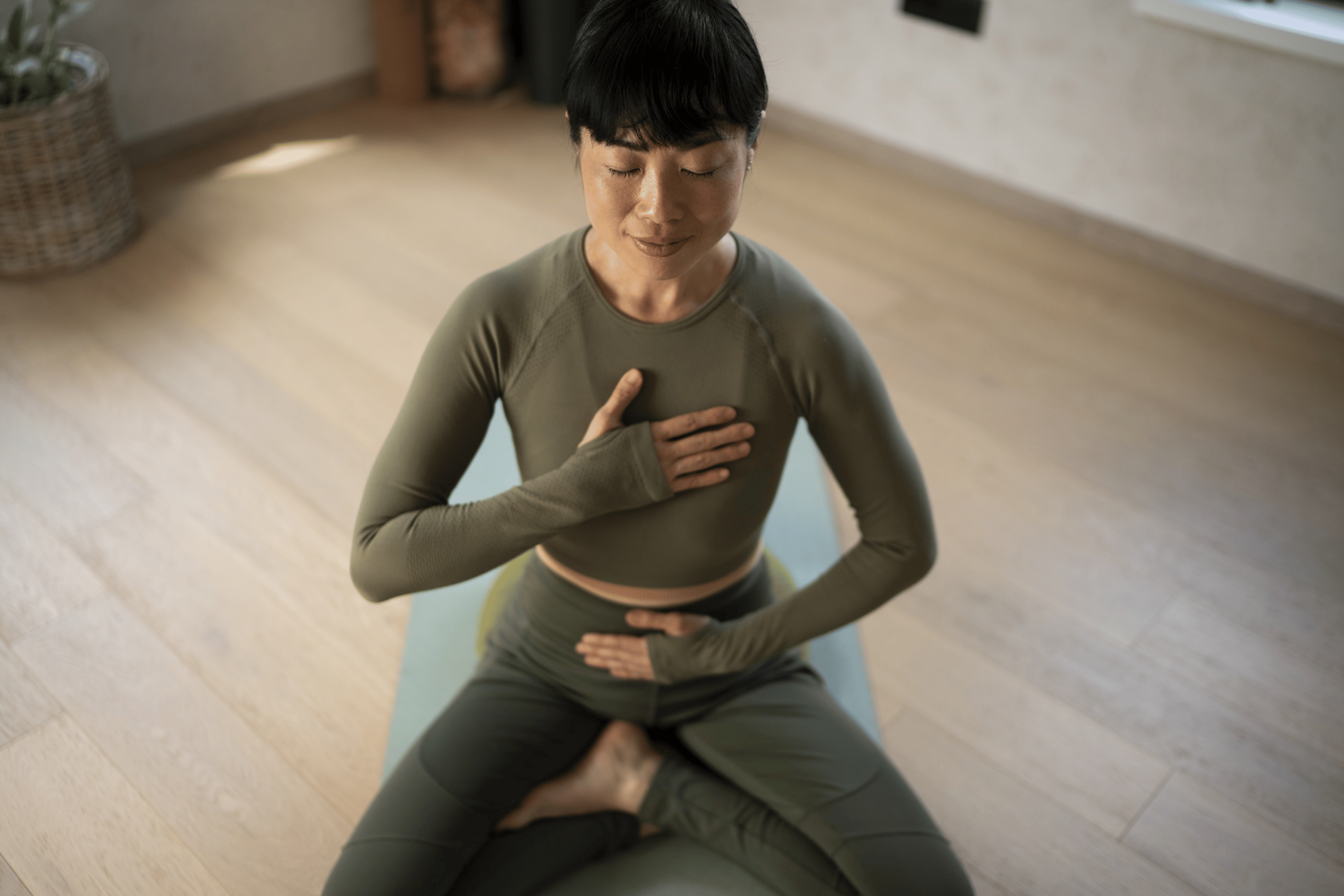 A woman sitting on a yoga mat and meditating while she takes off a week from working out. She is in an olive-green workout set and is sitting in lotus pose with her hands on her chest and belly.