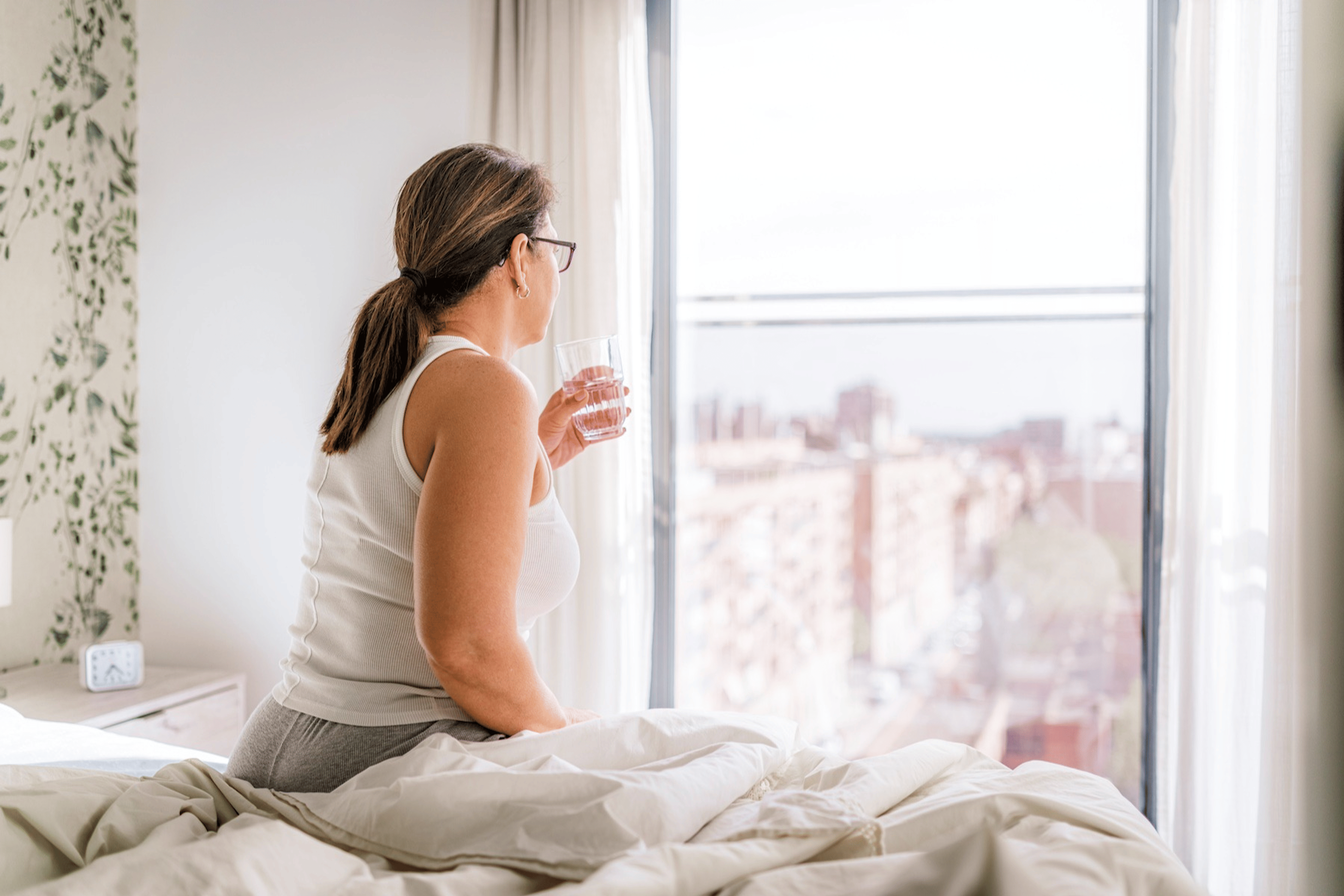 A woman sitting in bed drinking water first thing in the morning as she looks out the window.
