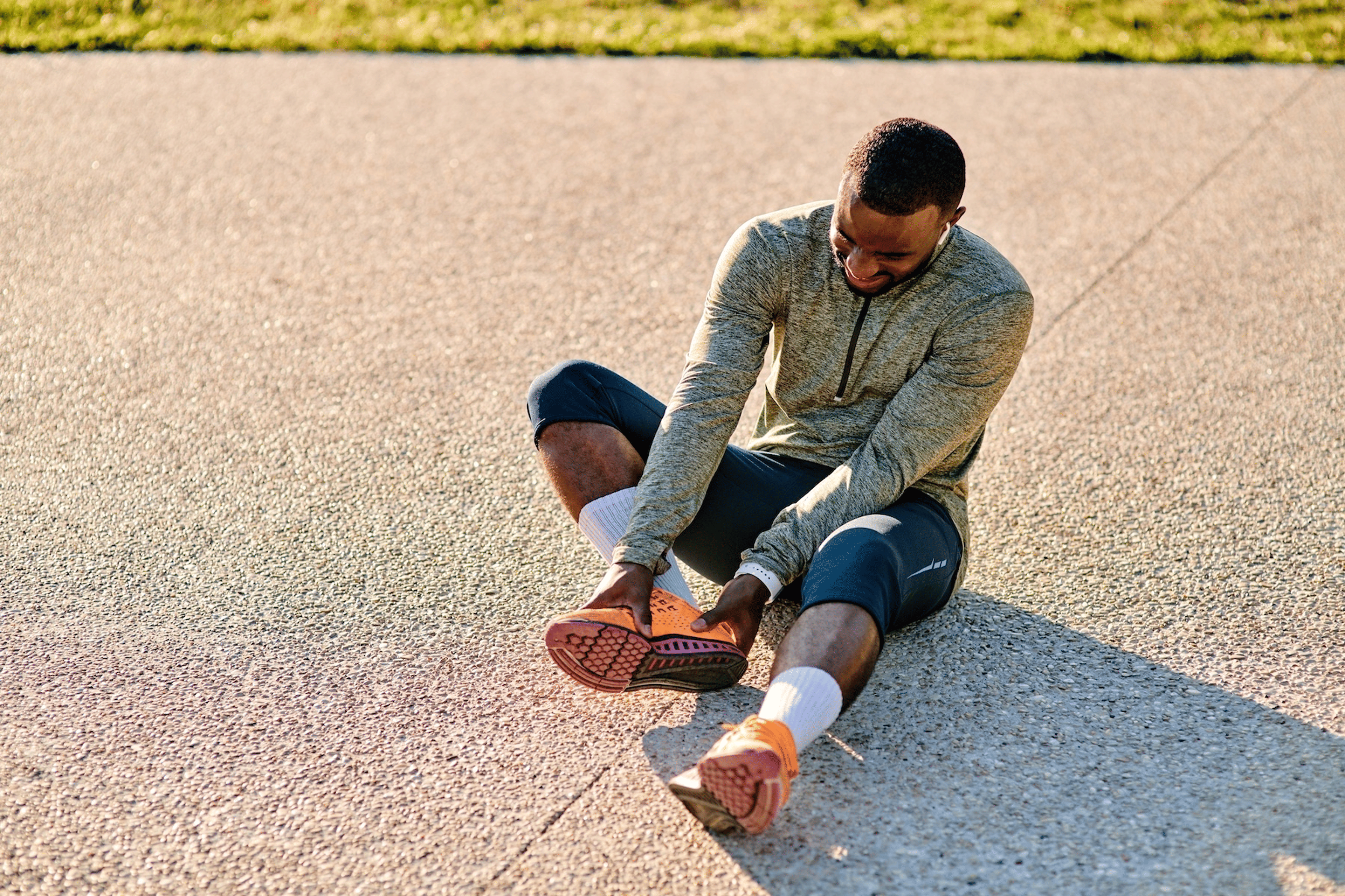A man in athletic clothes sitting down on the pavement while making a painful face as he takes off his running shoe. Learn how to prevent and treat blisters in this article.