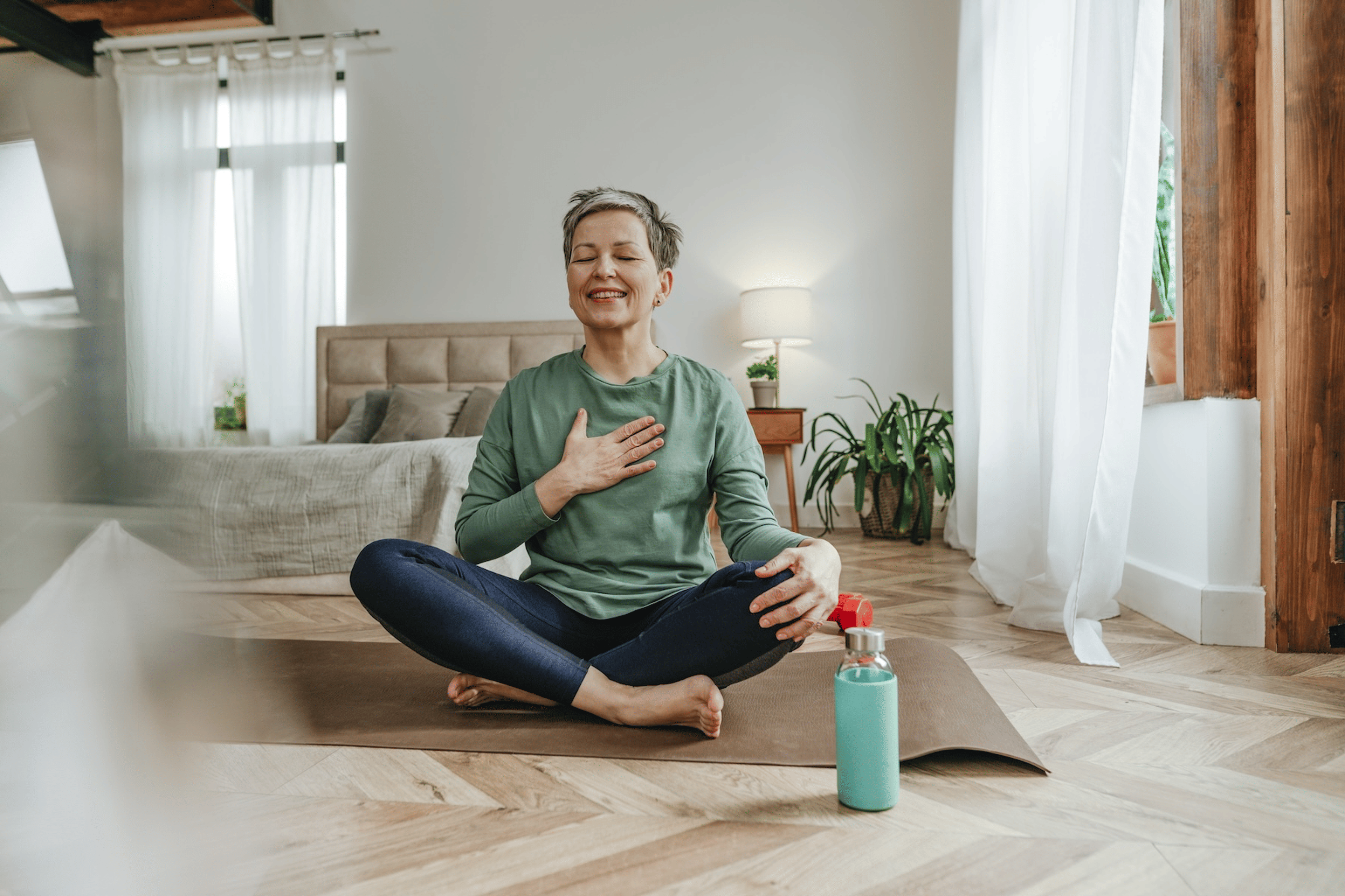 A woman meditating on an indoor yoga mat, with one hand on her chest and the other on her knee, to practice self-care during menopause.