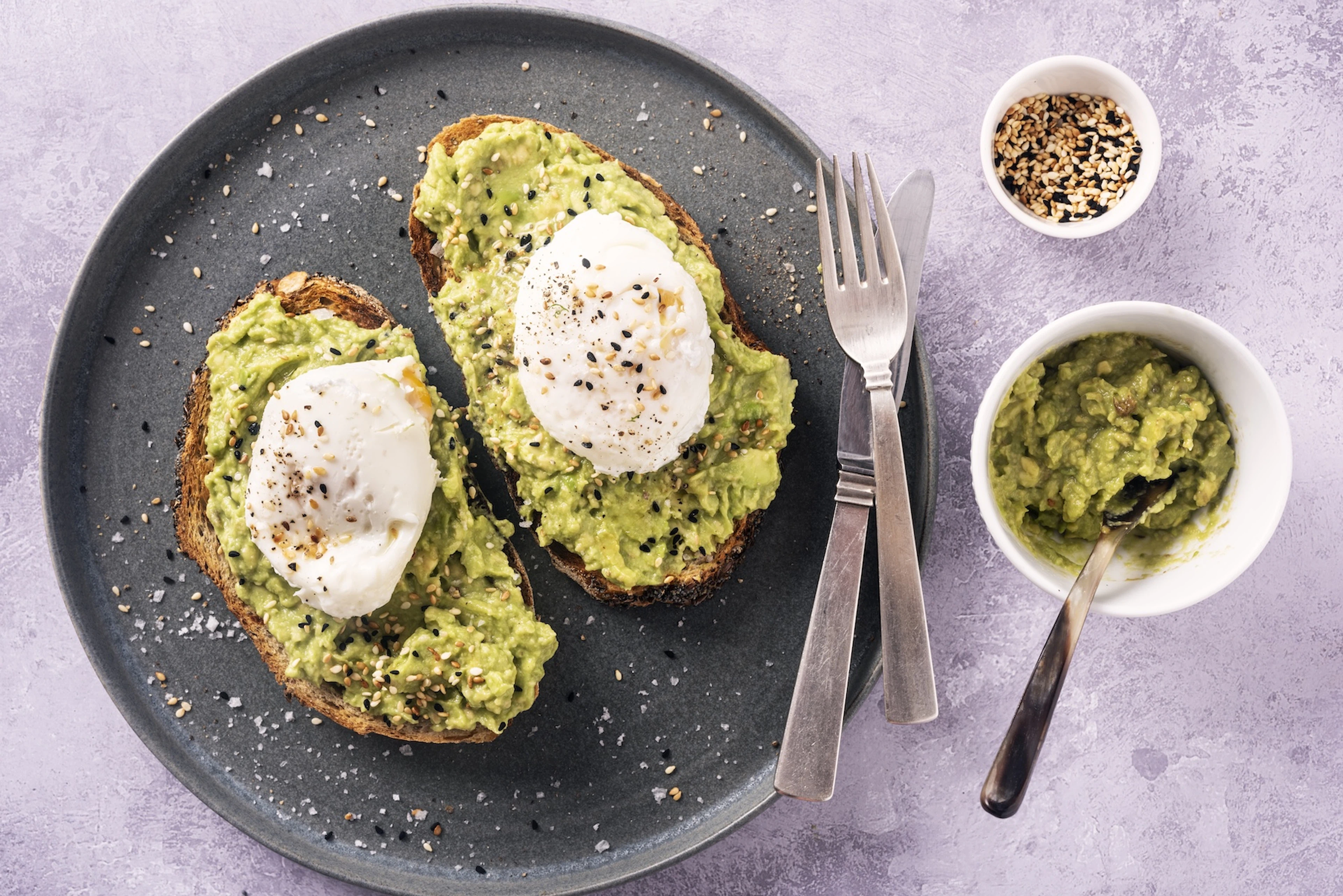 Two slices of avocado toast on a black plate. The toast is topped with poached eggs and everything seasoning. There are two bowls next to the plate,, one with mashed avocado and one with more seasoning.