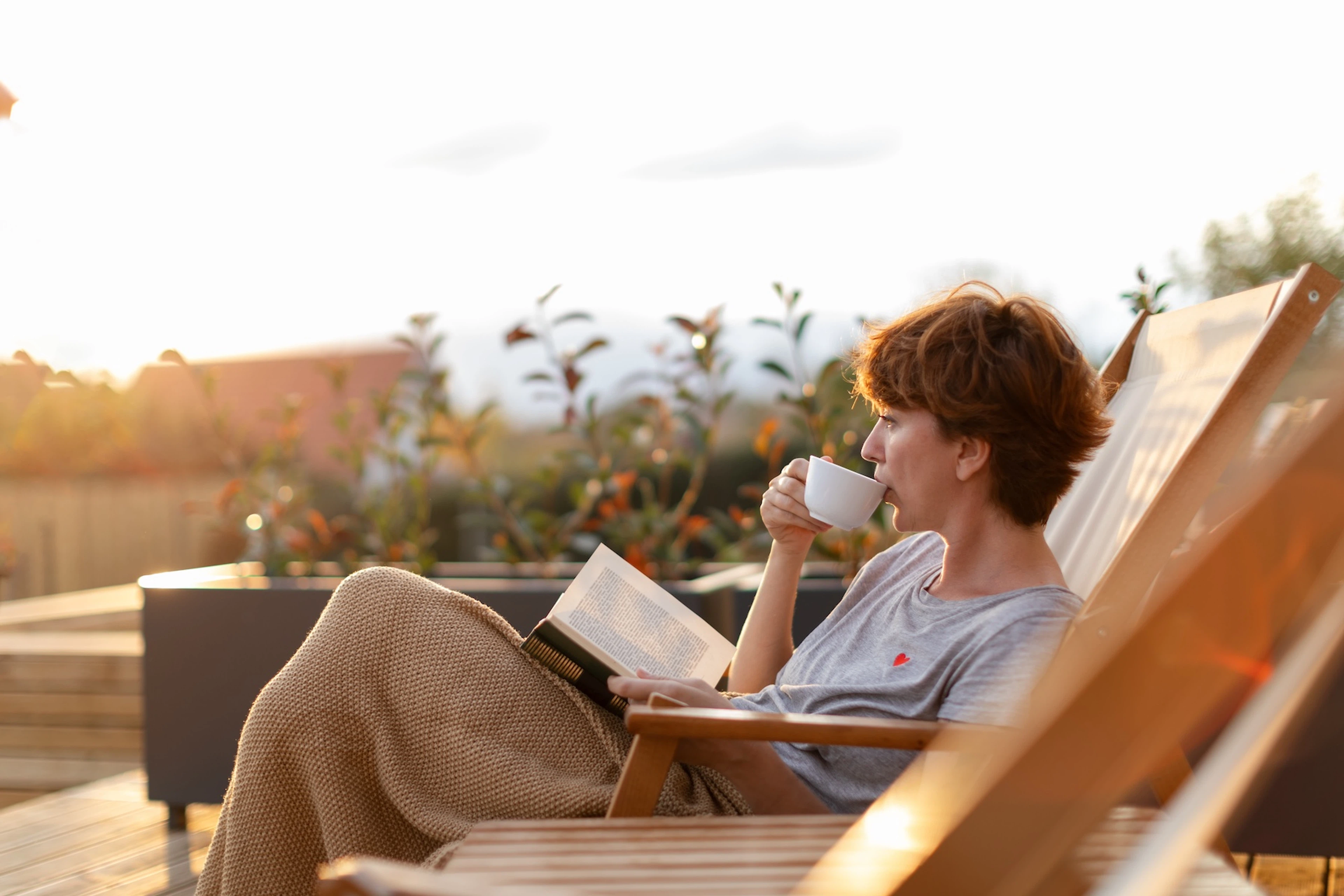 A woman sitting outside in the morning sunlight, reading a book, relaxing, and drinking coffee.