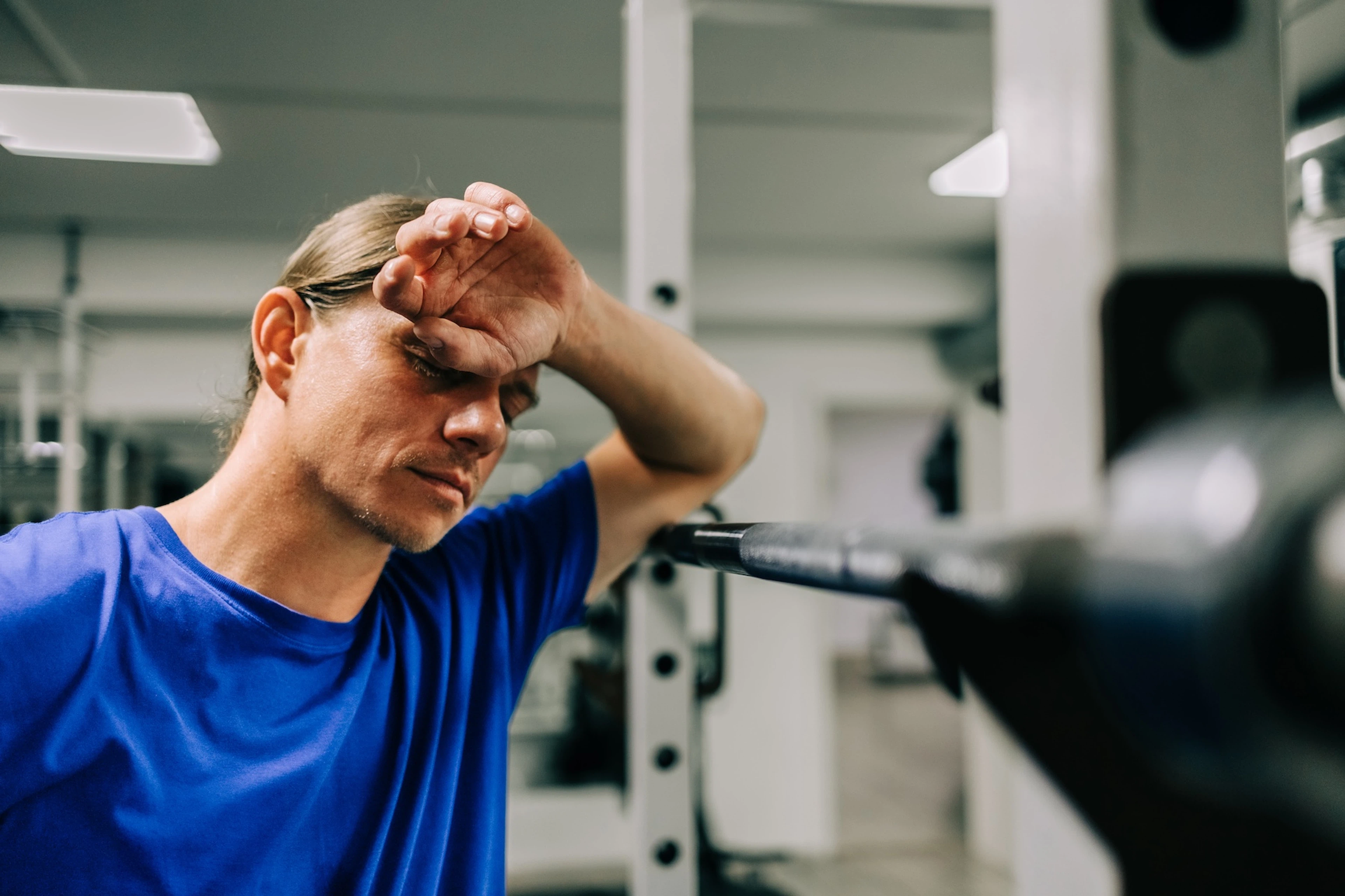 A tired man in the gym near a barbell. His hand is covering his face and his eyes are closed. He's wearing a bright blue shirt.