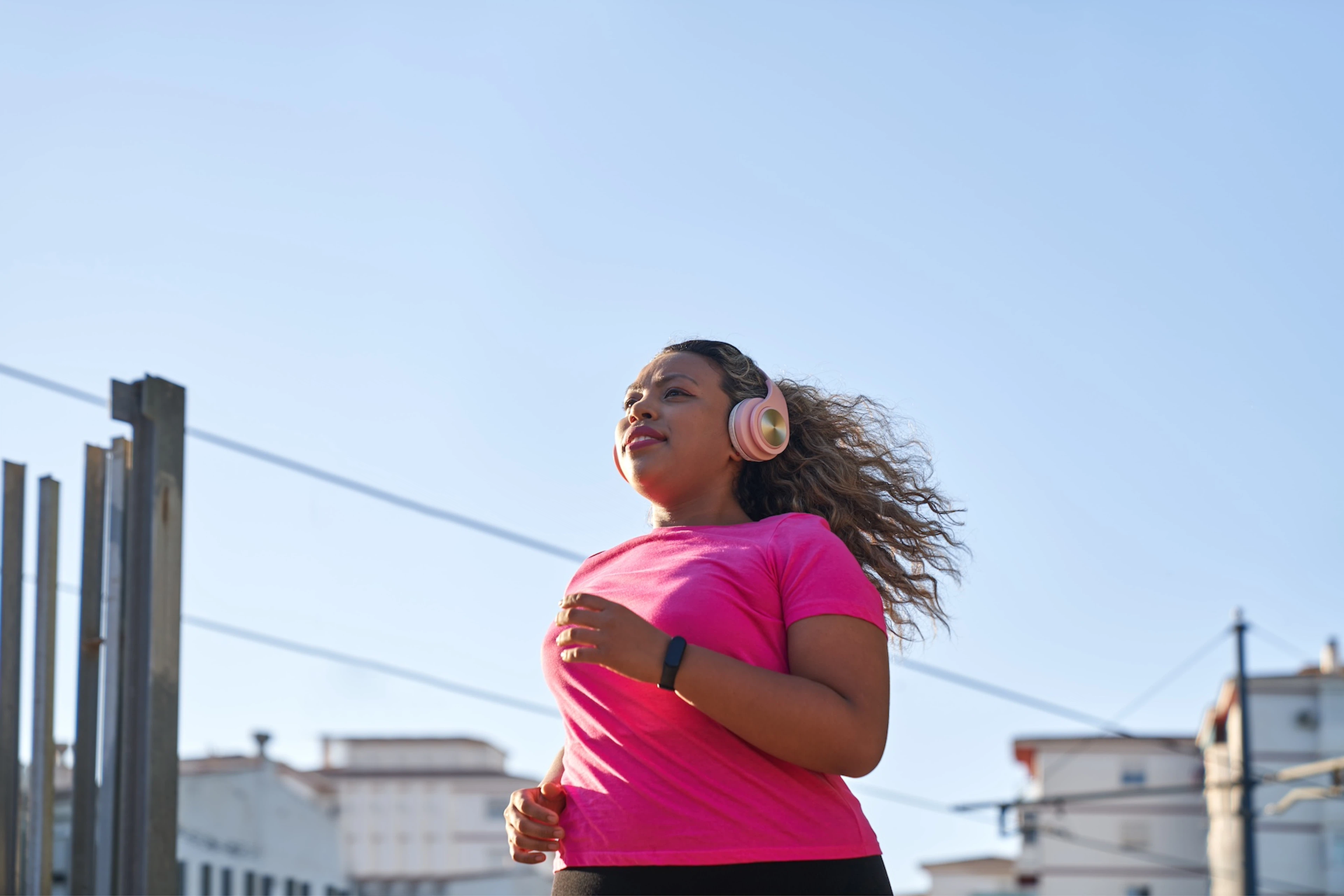 A woman in a pink shirt and headphones running outside through a city. Learn how to motivate yourself to work out in this article.