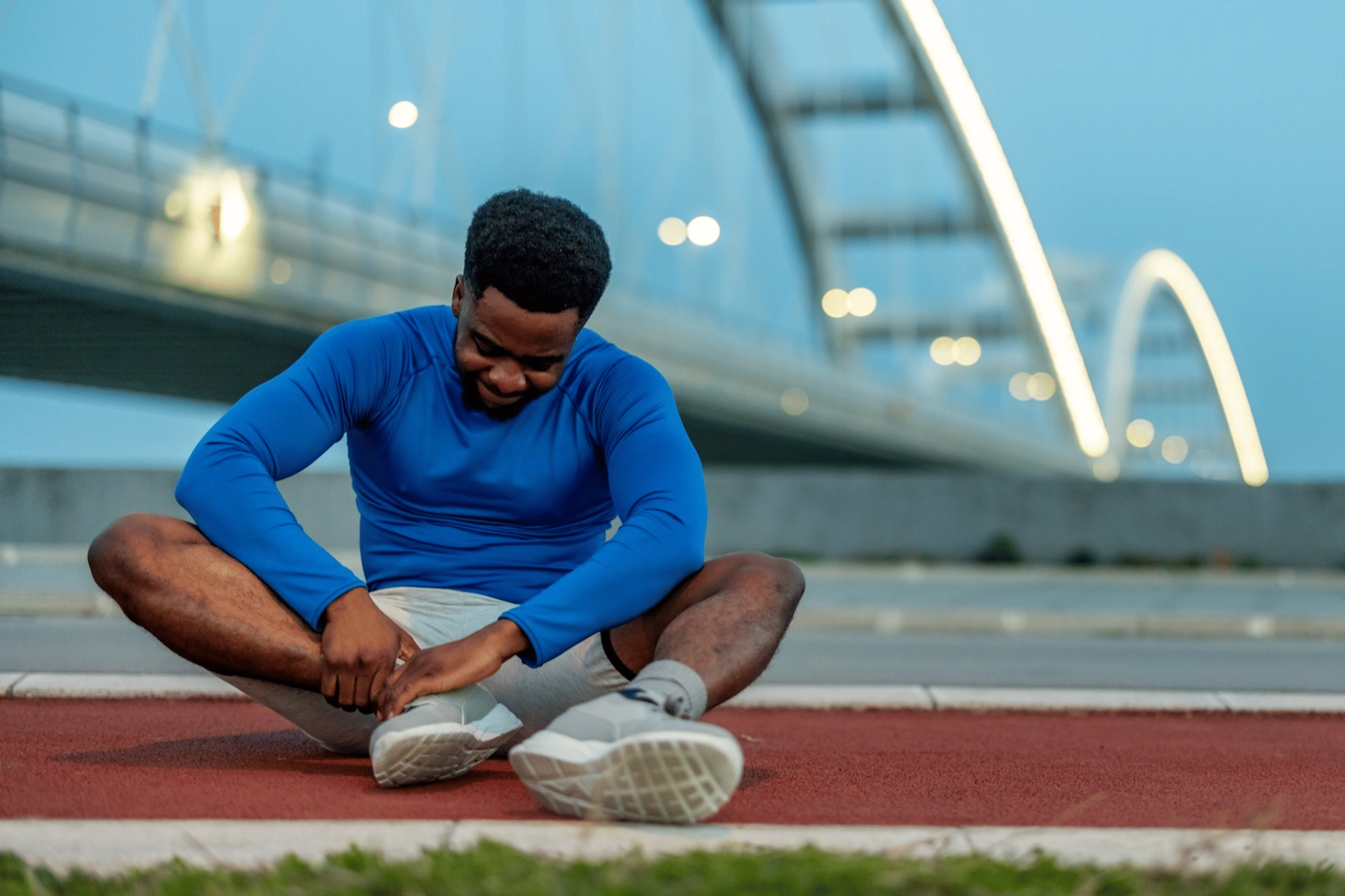 A male athlete sitting on the ground and holding his right ankle. He's experiencing ankle pain after running.