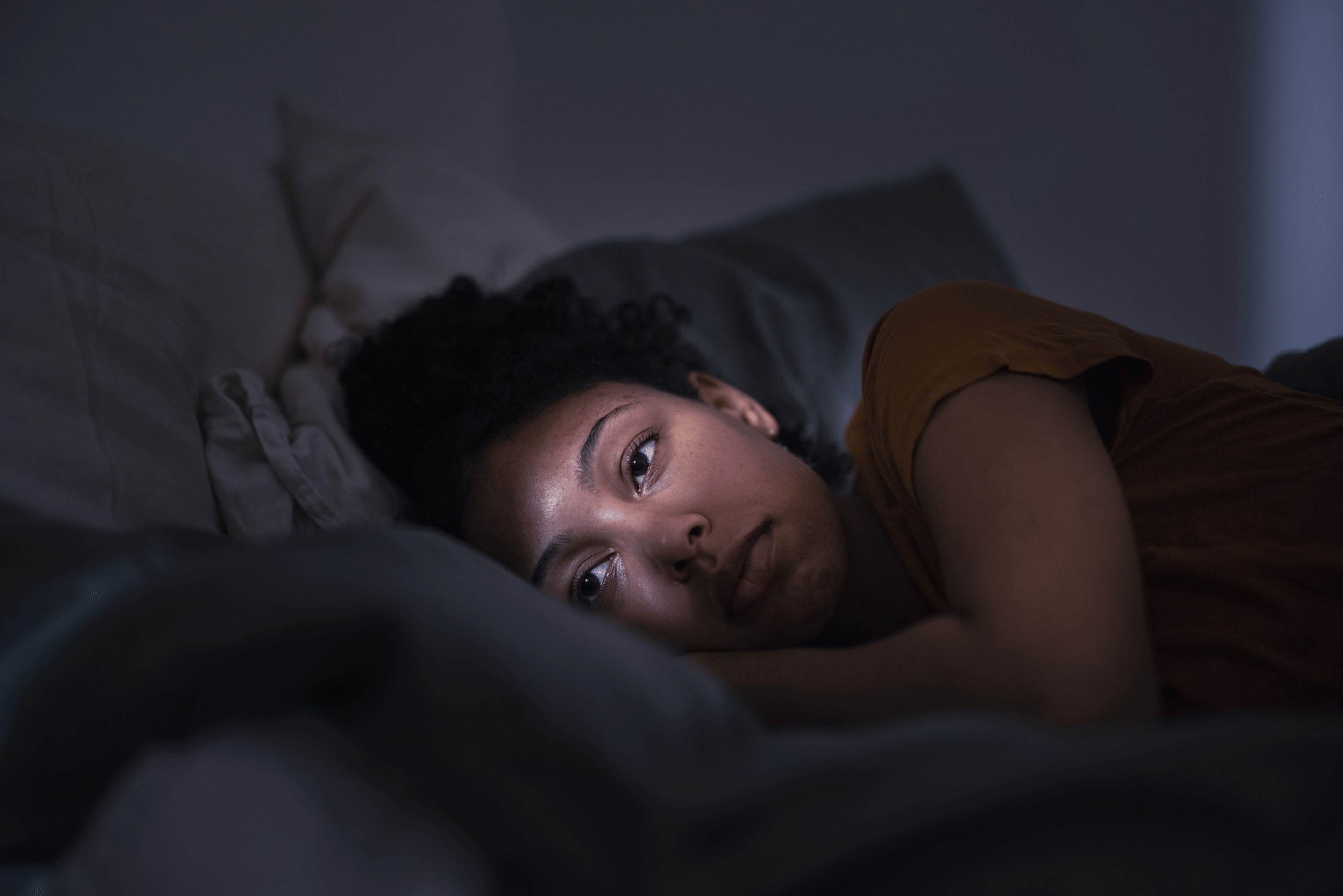 A woman experiencing anxiety at night. She's lying in bed on her side with her eyes open, staring off into space.