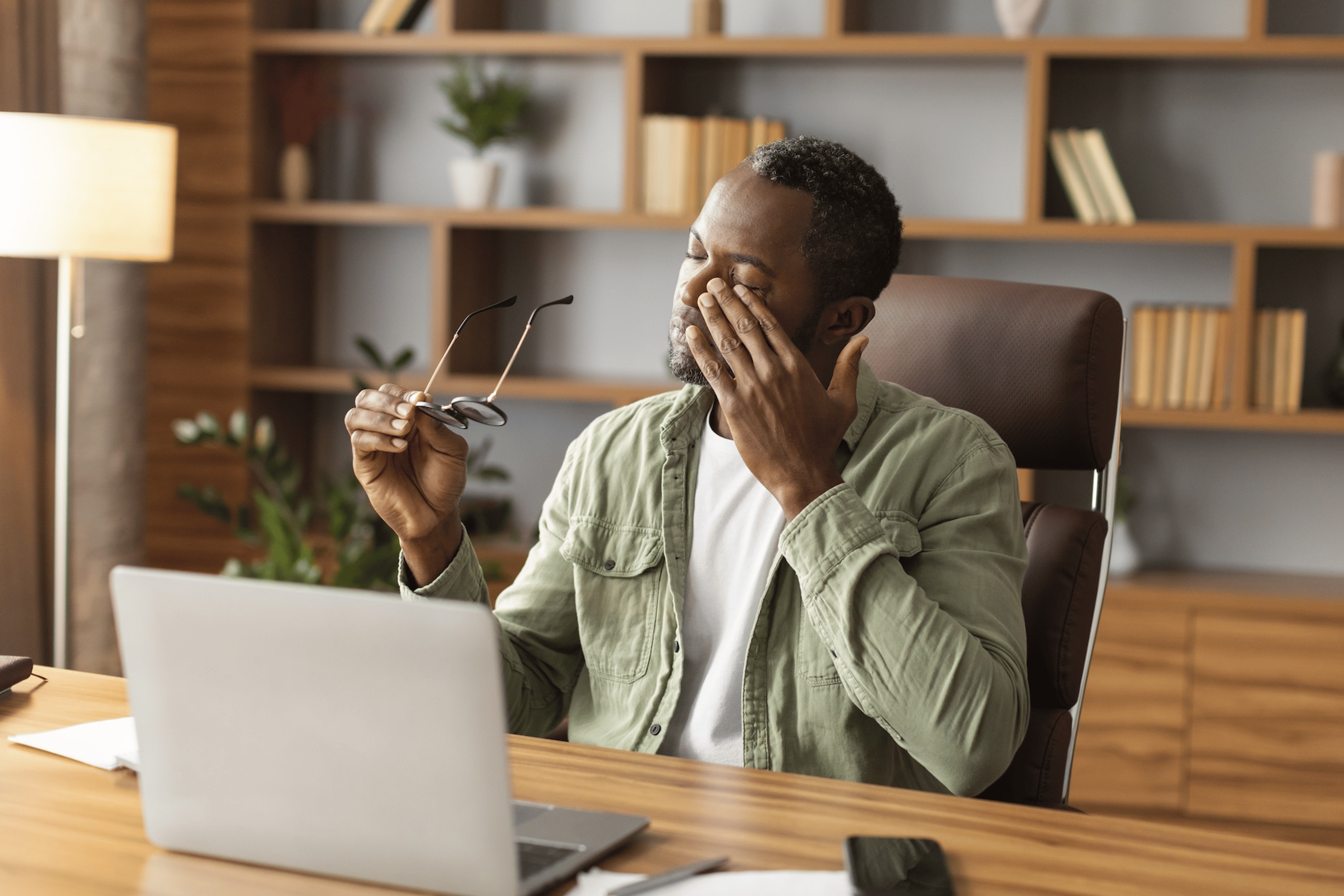 An overtired man sitting at his desk rubbing his eye while holding his glasses in his hand.