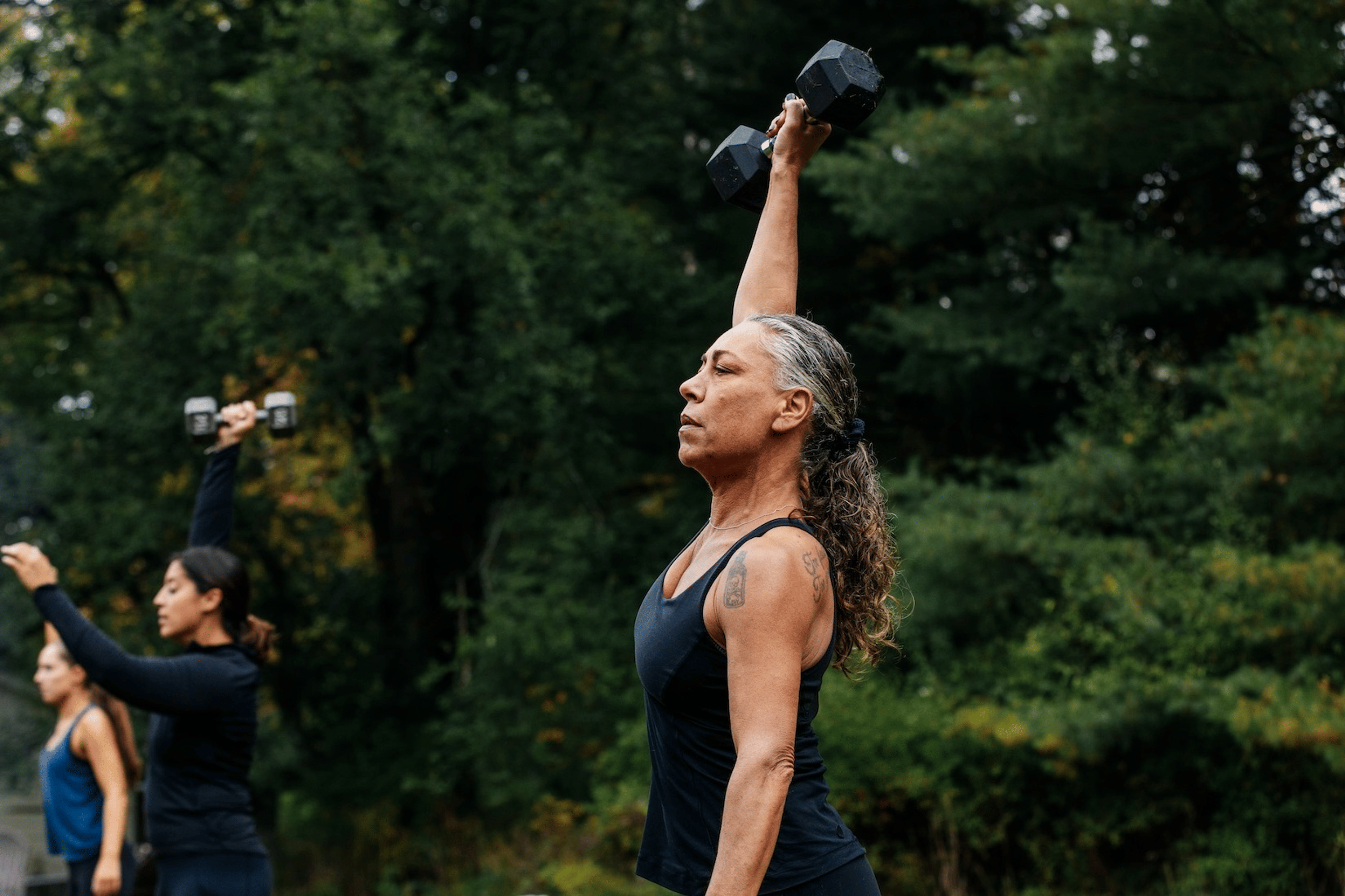 A middle-aged woman lifting a dumbbell in the air while doing an outdoor workout.