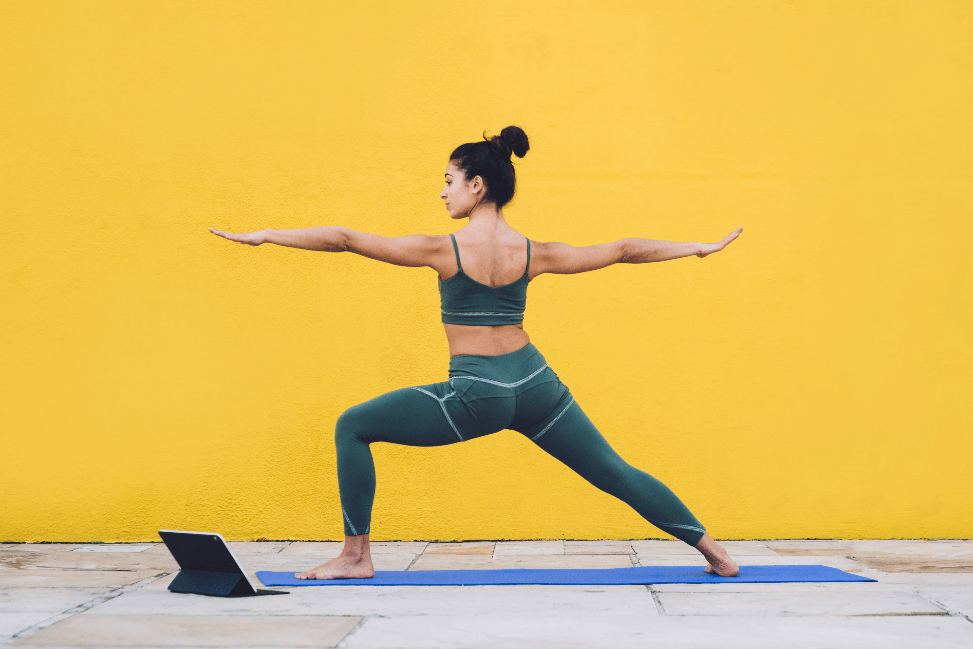 Side view of focused calm female in sportive outfit concentrating in warrior pose on yoga mat with yellow background behind