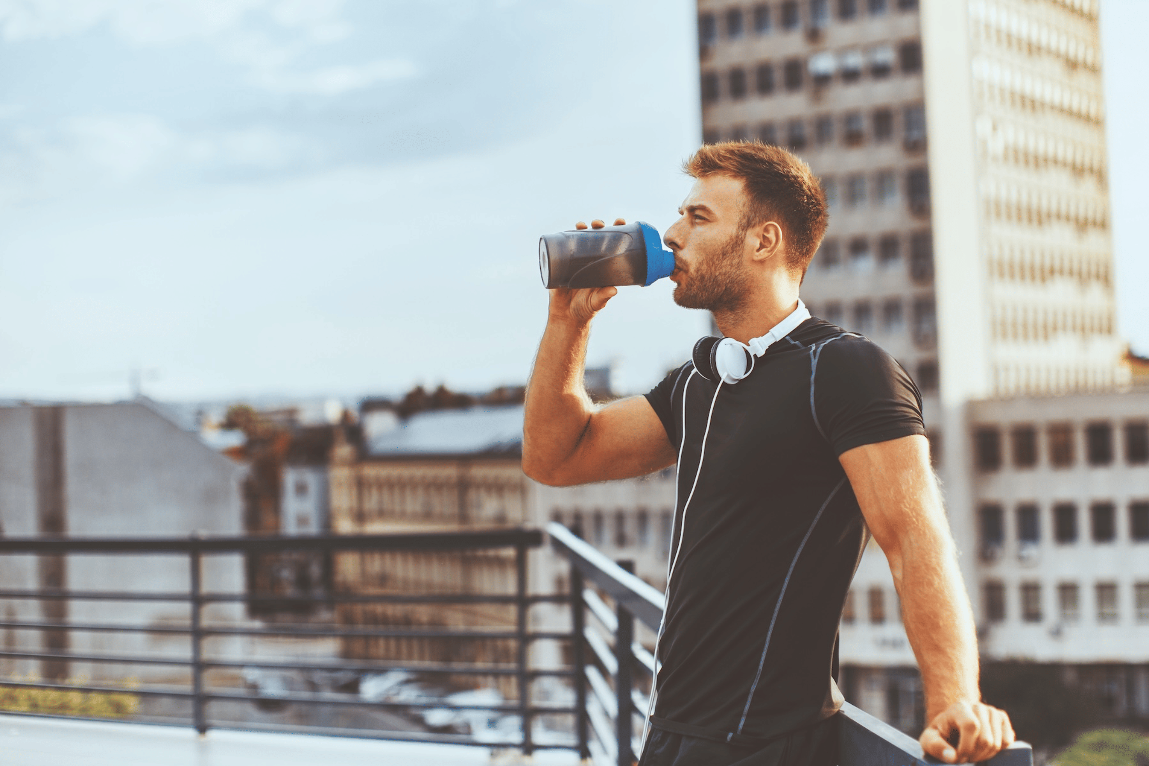 A man drinking creatine from a shaker bottle. He's outside and leaning against a fence after a workout.