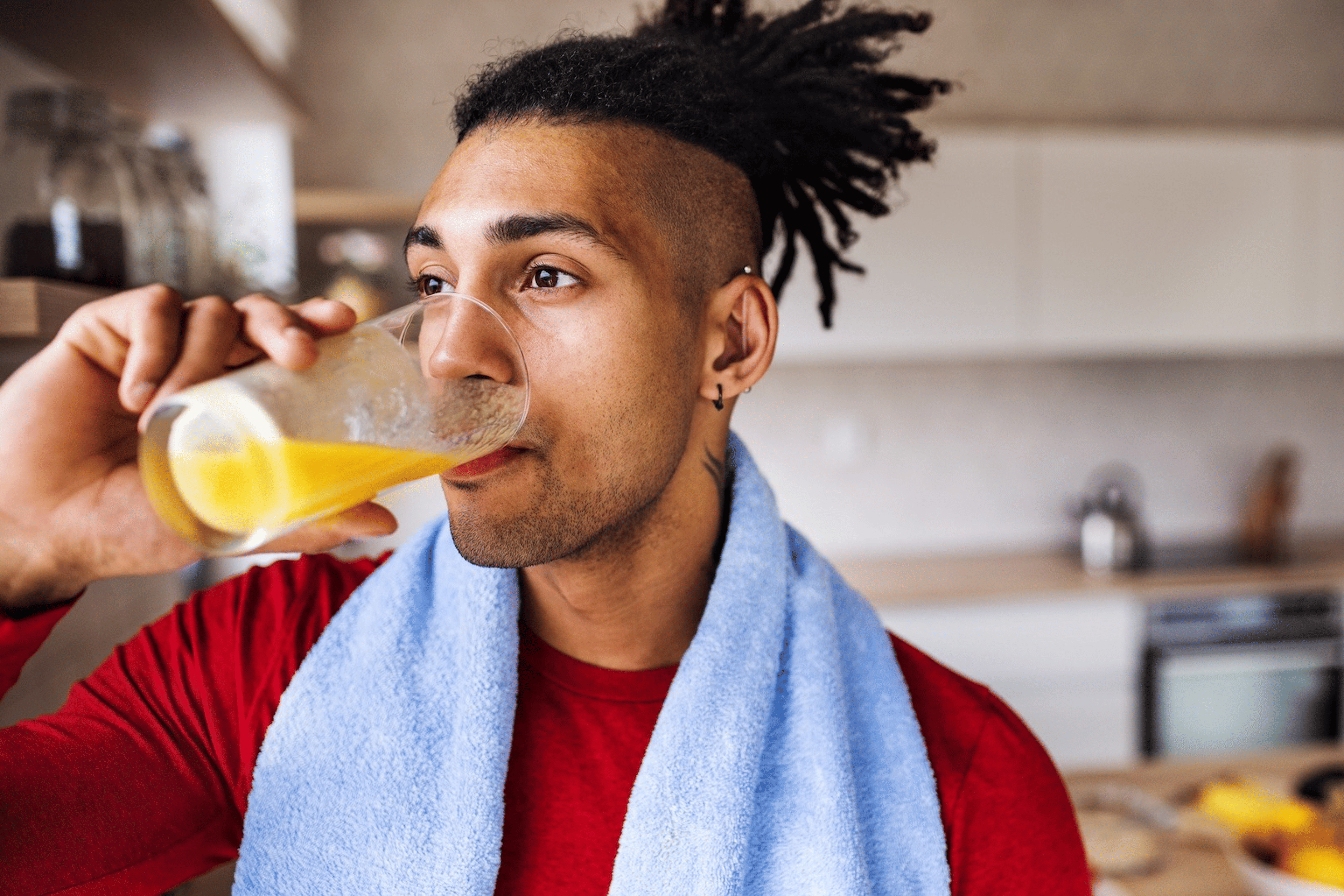 An athlete drinking a glass of orange juice before going on a run. He's in his kitchen and has a towel around his neck.