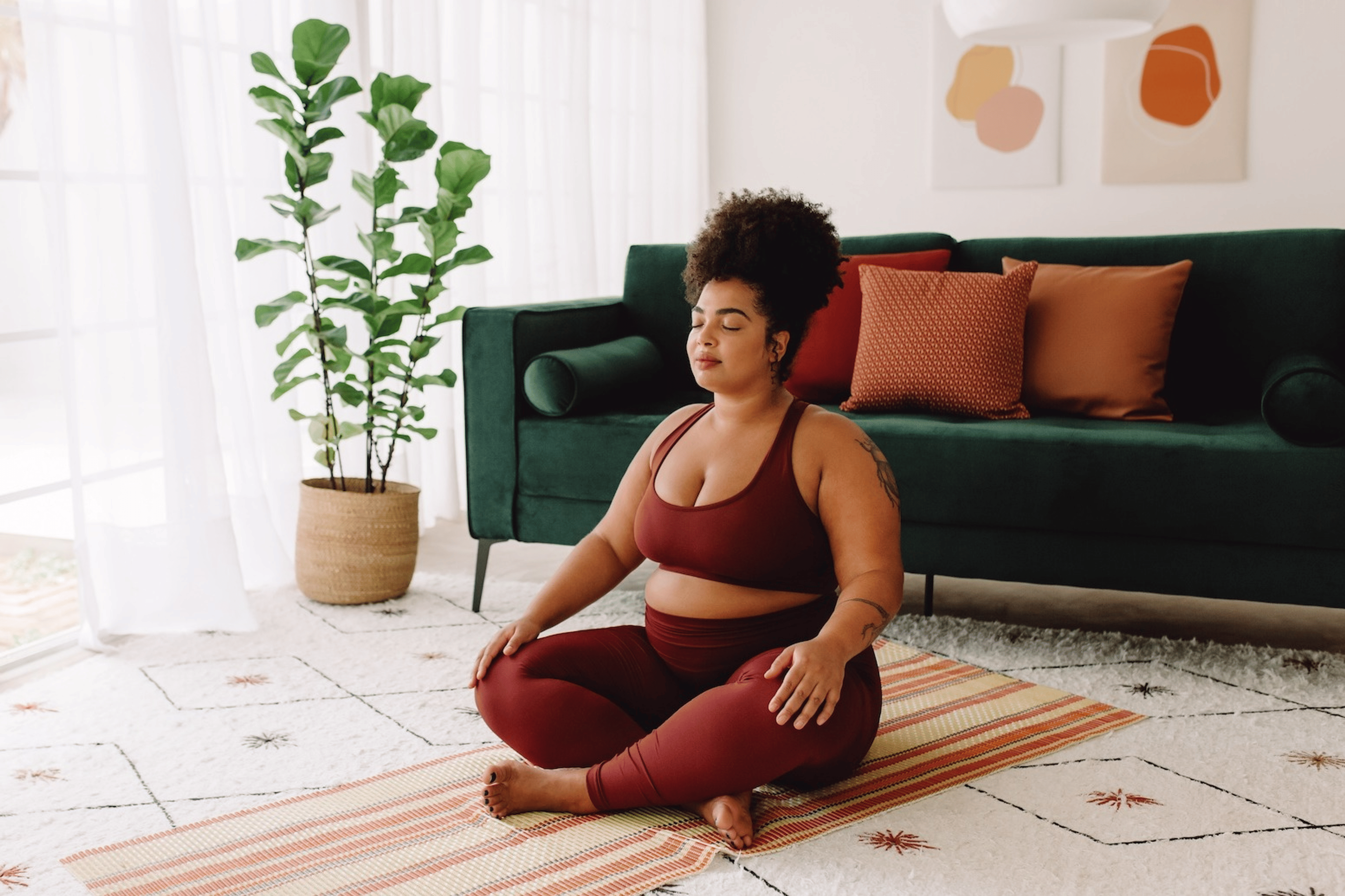 A woman practicing healthy habits by meditating at home. She's wearing activewear and sitting on a blanket on the floor in a sunny living room.