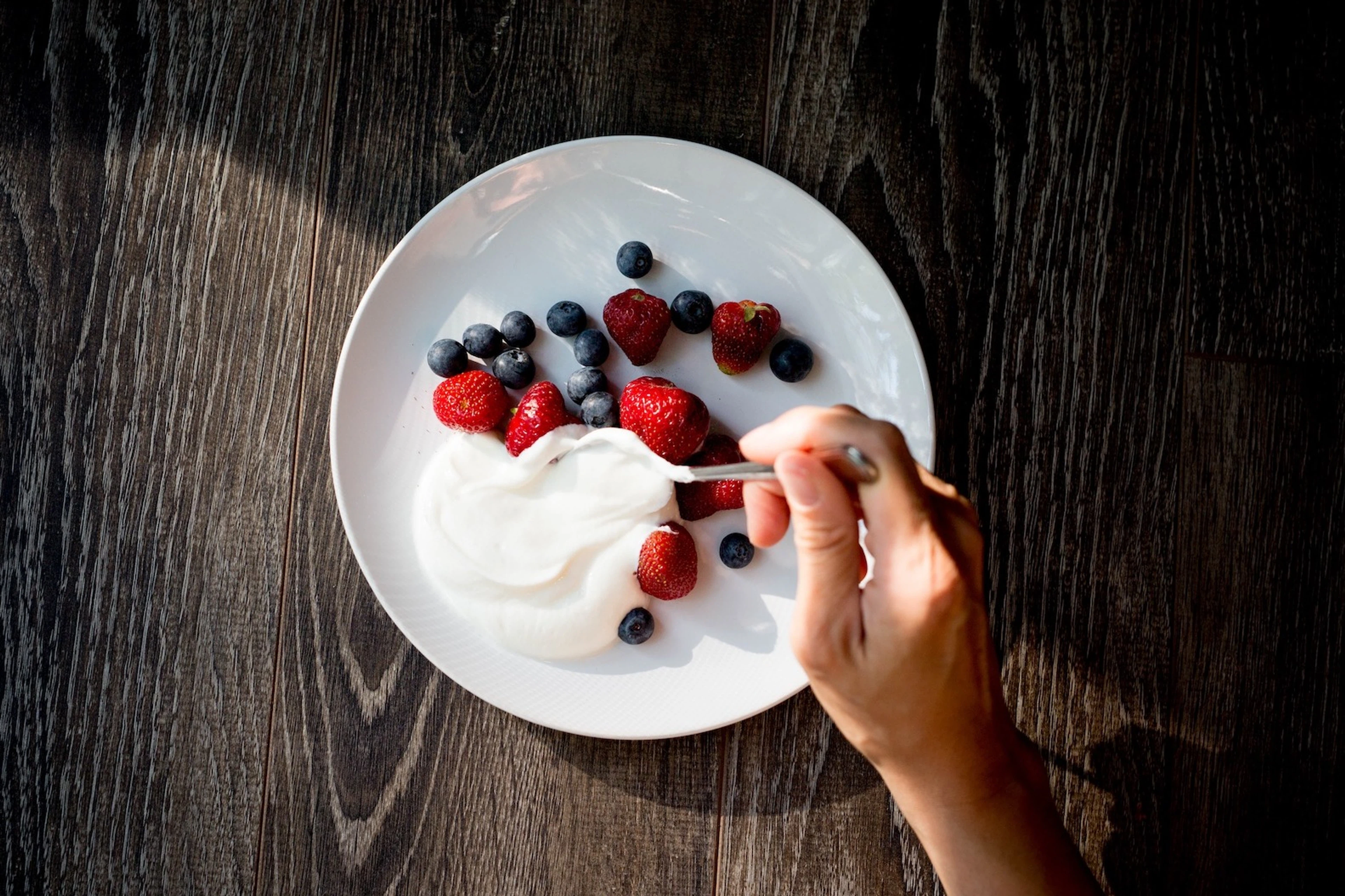 Close-up photo of a woman spooning Greek yogurt onto a white plate with blueberries and strawberries.