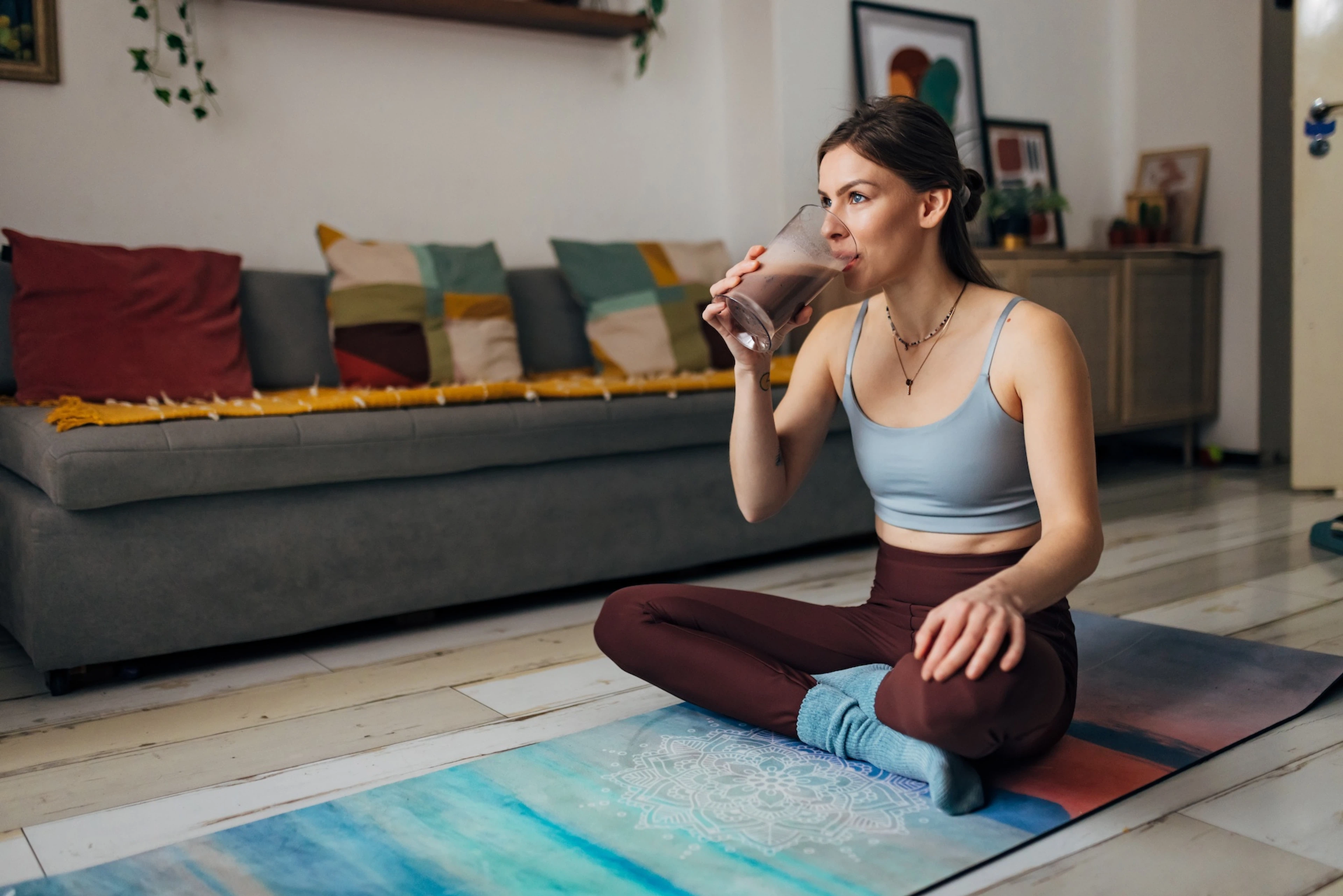 A millennial woman sitting on a yoga mat at home and drinking a glass of chocolate milk after a workout.