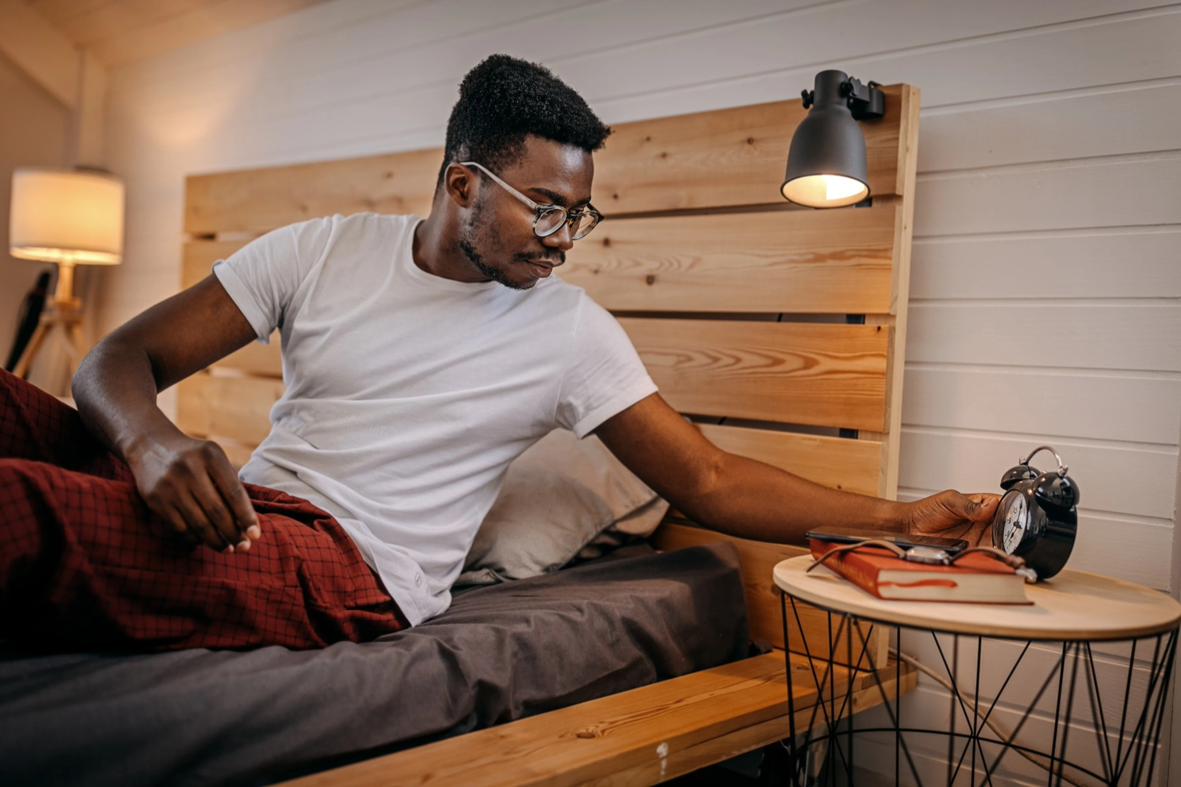 A man going to bed, looking at his alarm clock while sitting under the covers.