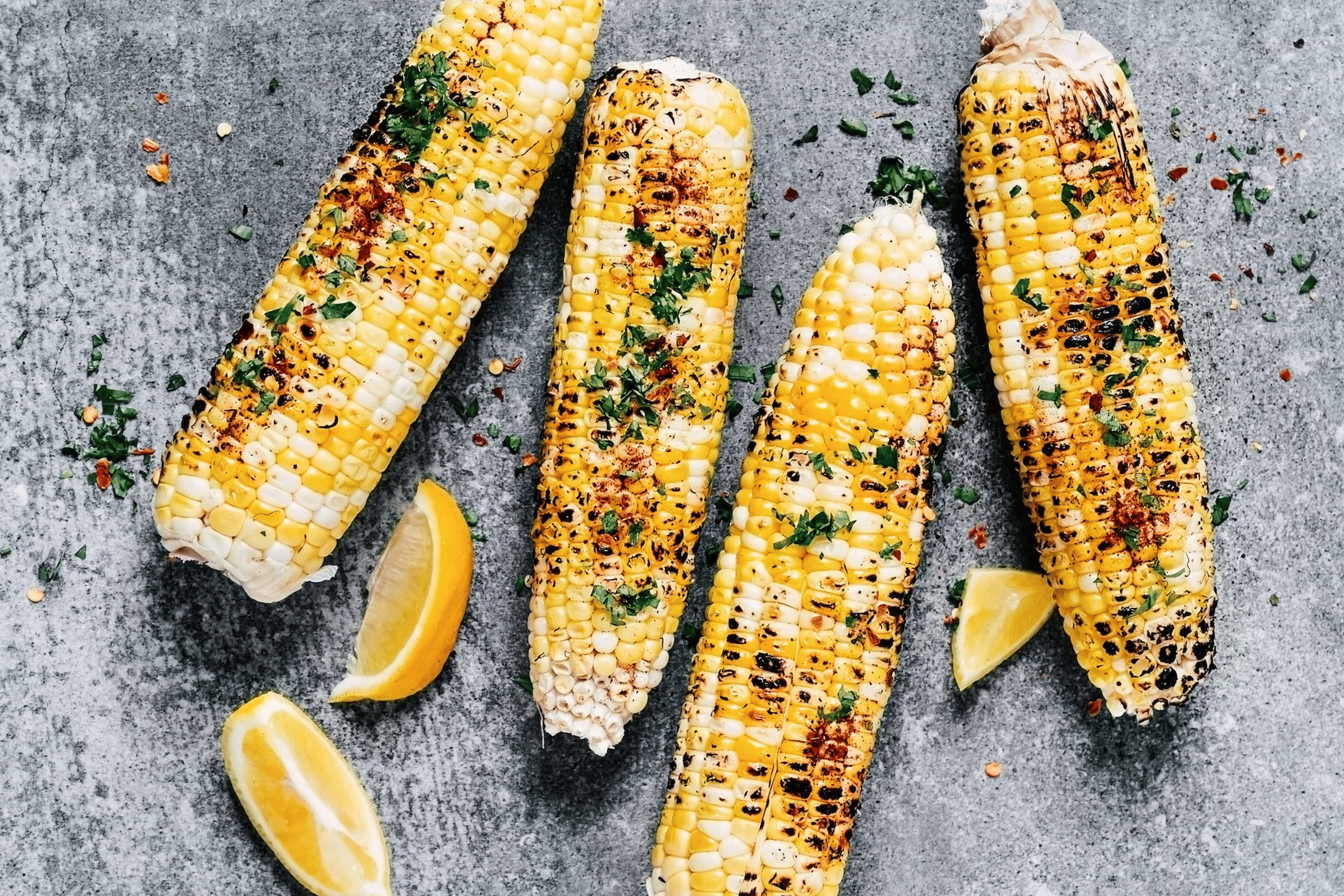 Four grilled cobs of corn with fresh herbs resting on a gray surface next to lemon slices. Corn is a high-protein vegetable.