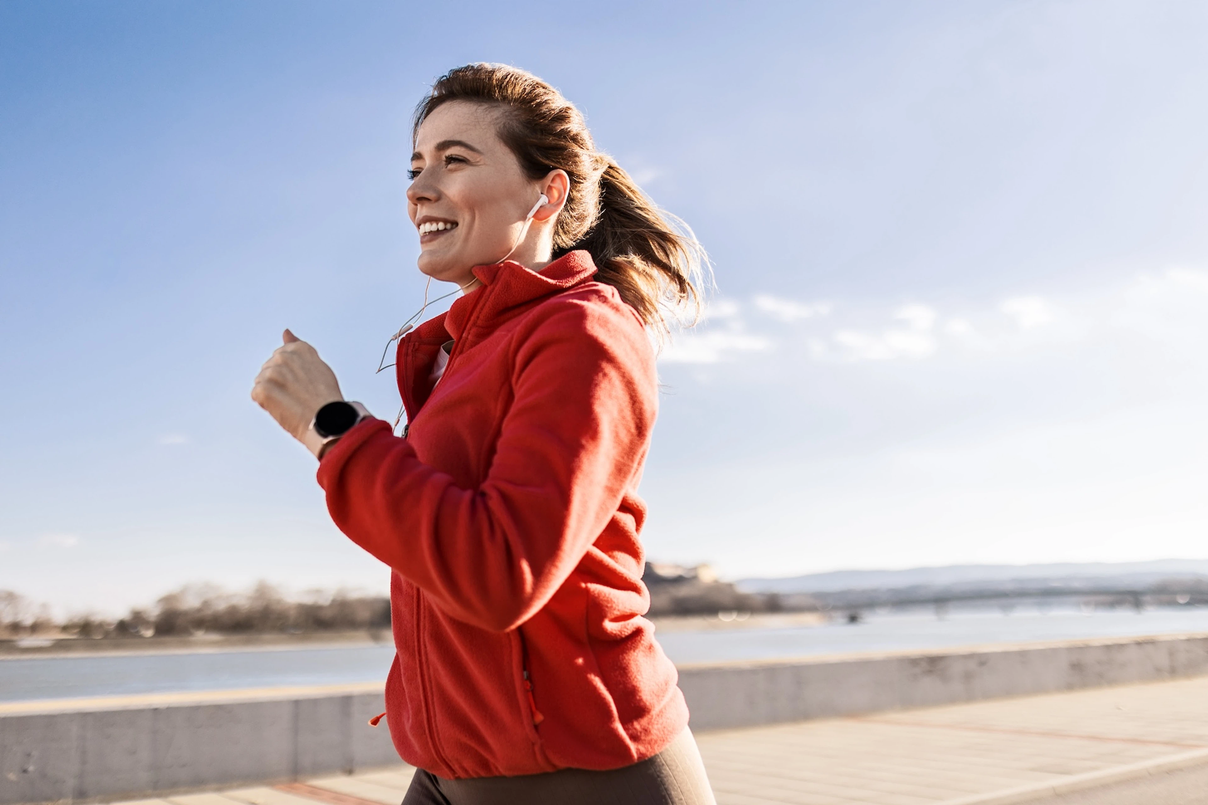 A happy woman going a run outside. She's wearing a red zip-up hoodie.