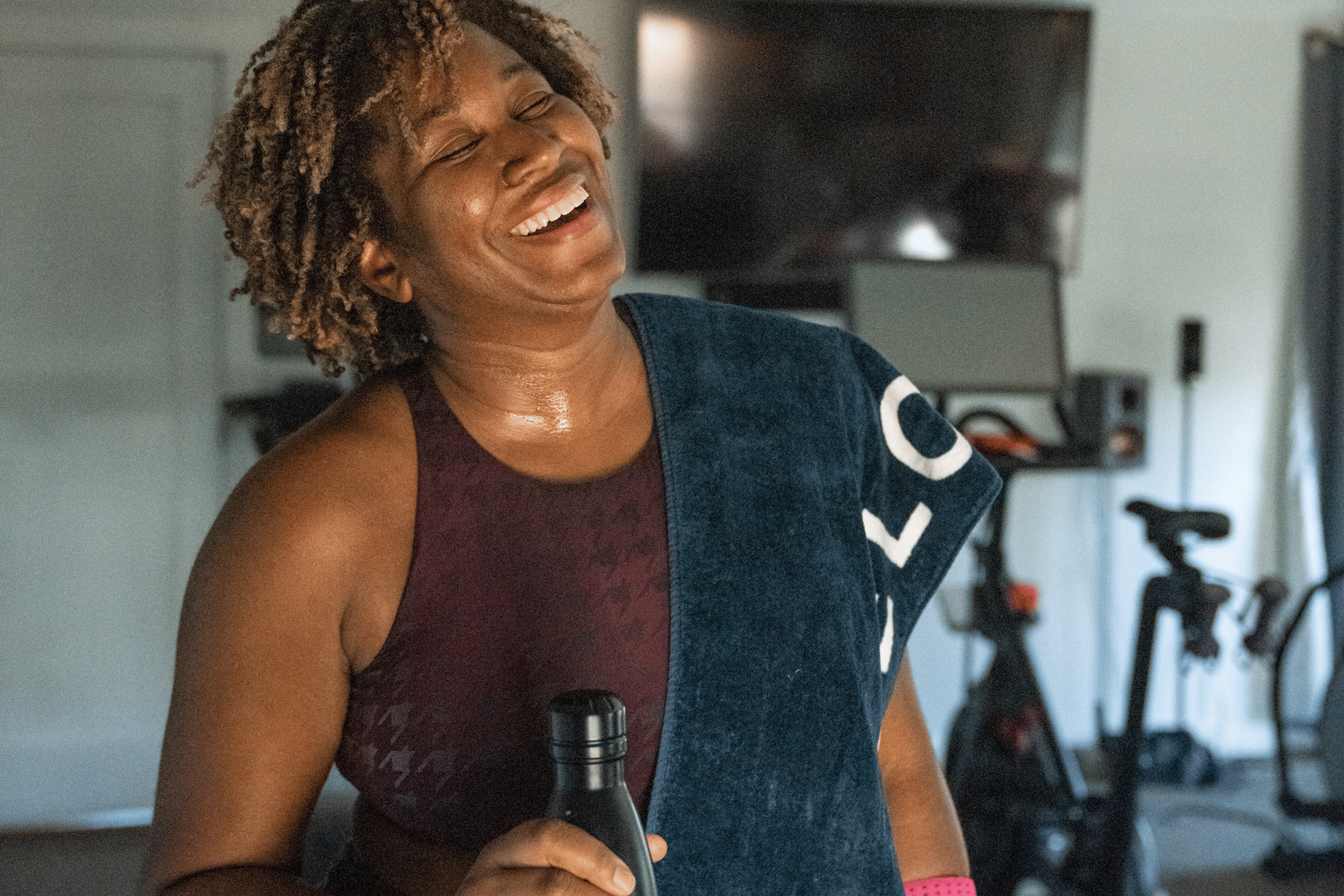 A woman laughing and smiling after a workout. She is holding a water bottle and has a towel draped around her left shoulder.