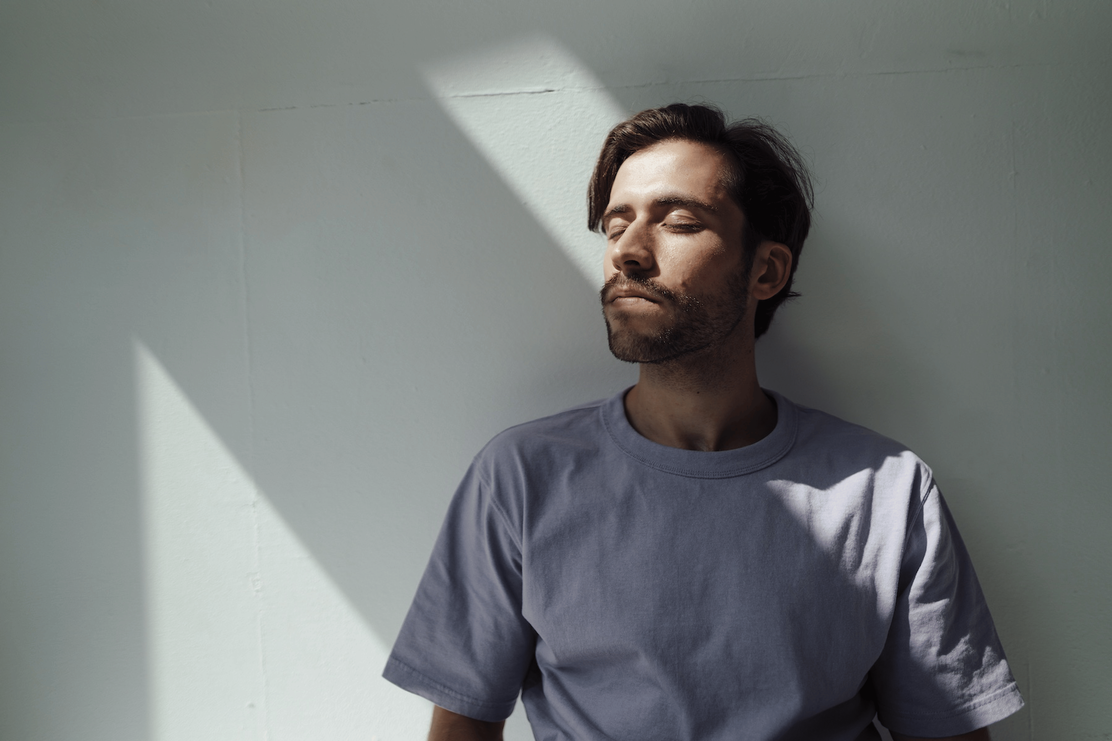 A relaxed young man leaning against a sunny white wall while closing his eyes and practicing ujjayi breathing.