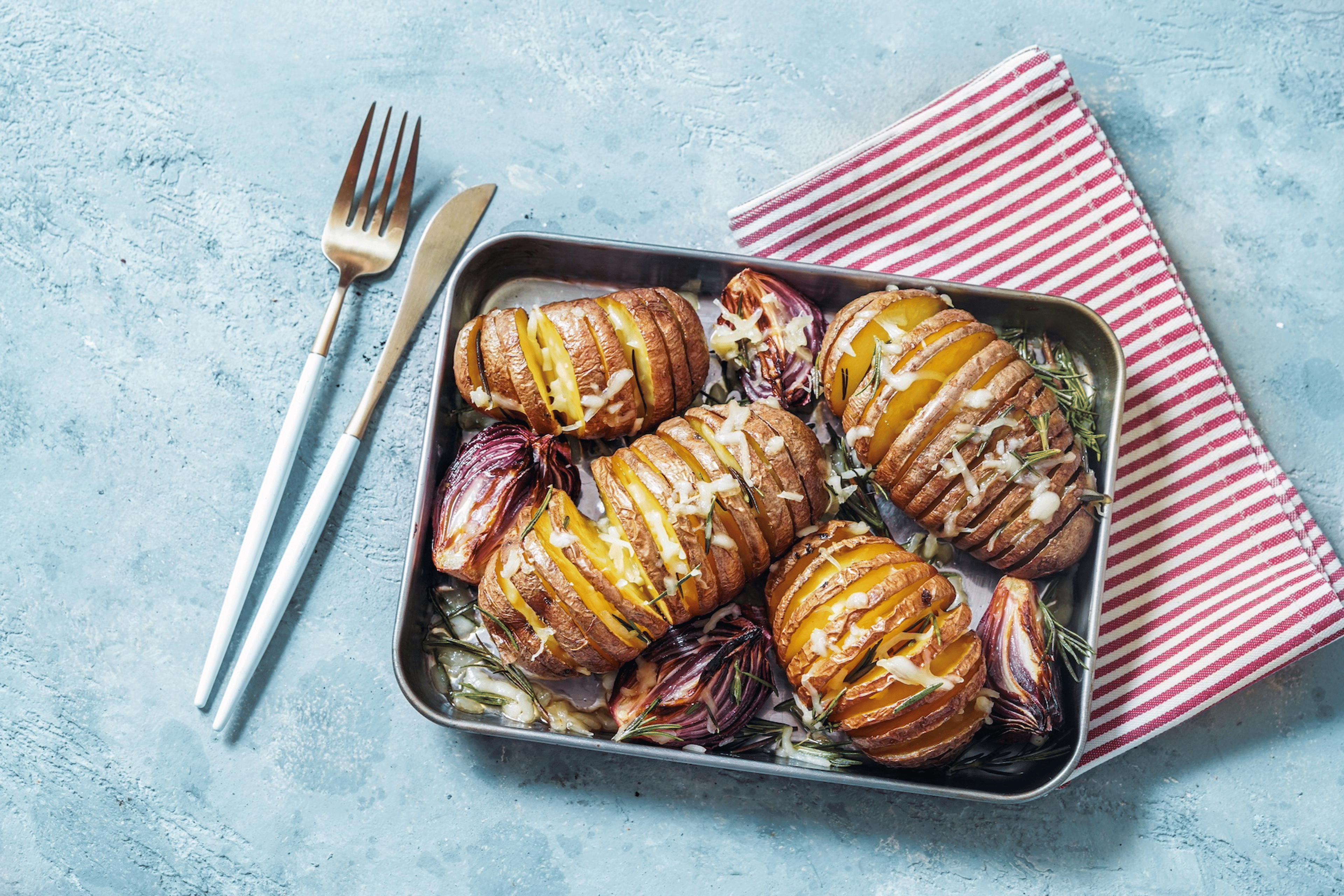 A tray with hasselback potatoes on it next to a striped napkin and cutlery. Baked potatoes are high-protein vegetables.
