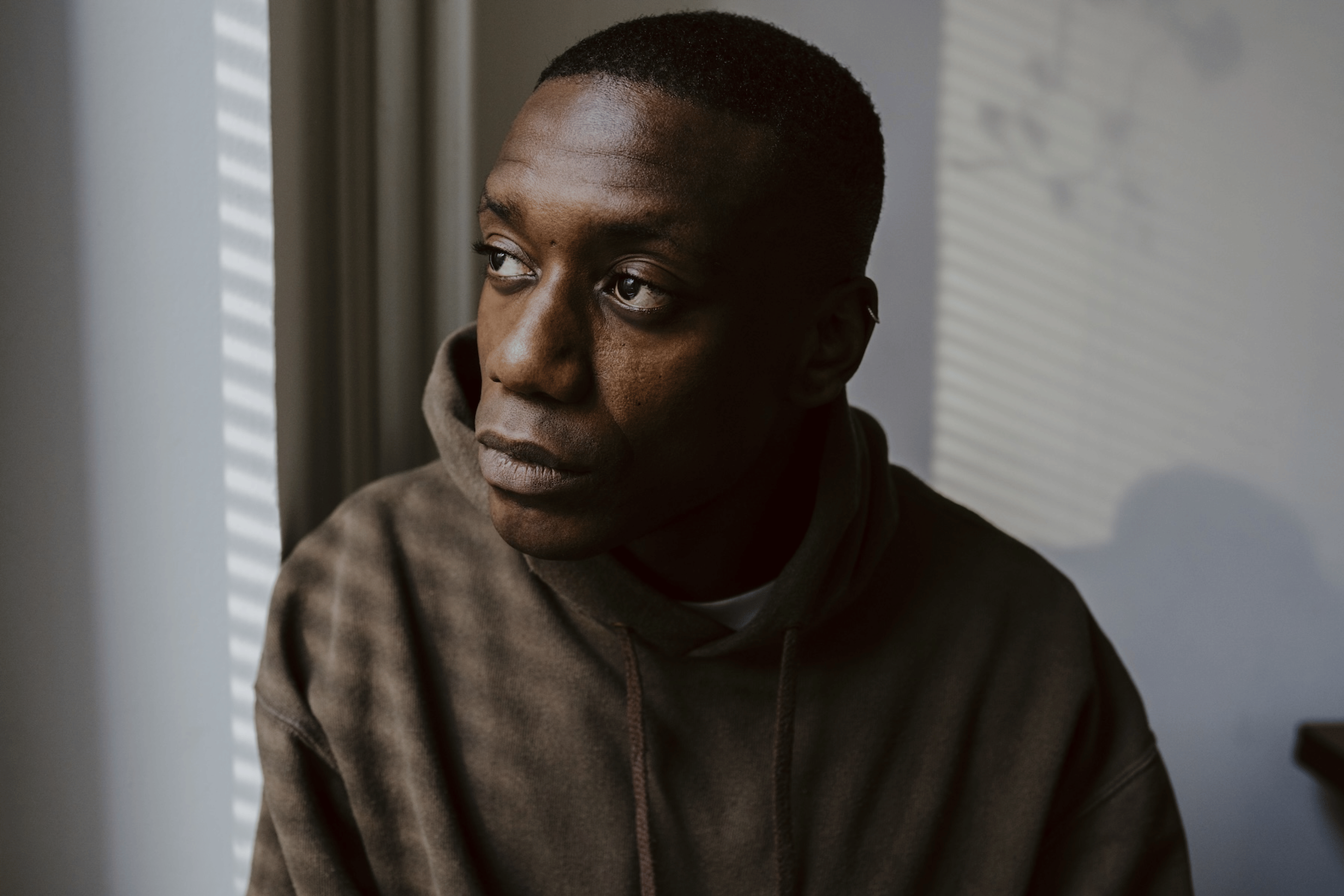 A man looking out a window. He is wearing a brown hoodie and is sick from stress.