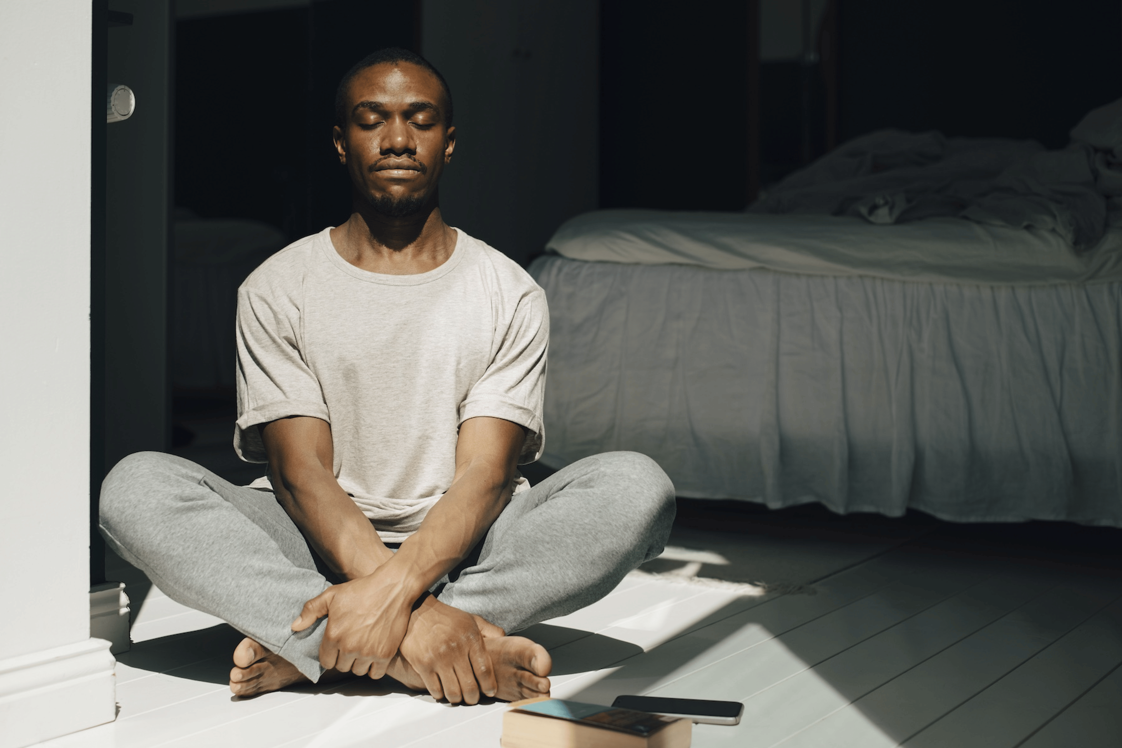 A man calmly sitting on the floor and meditating in his bedroom. Check out eight meditation tips in this article.