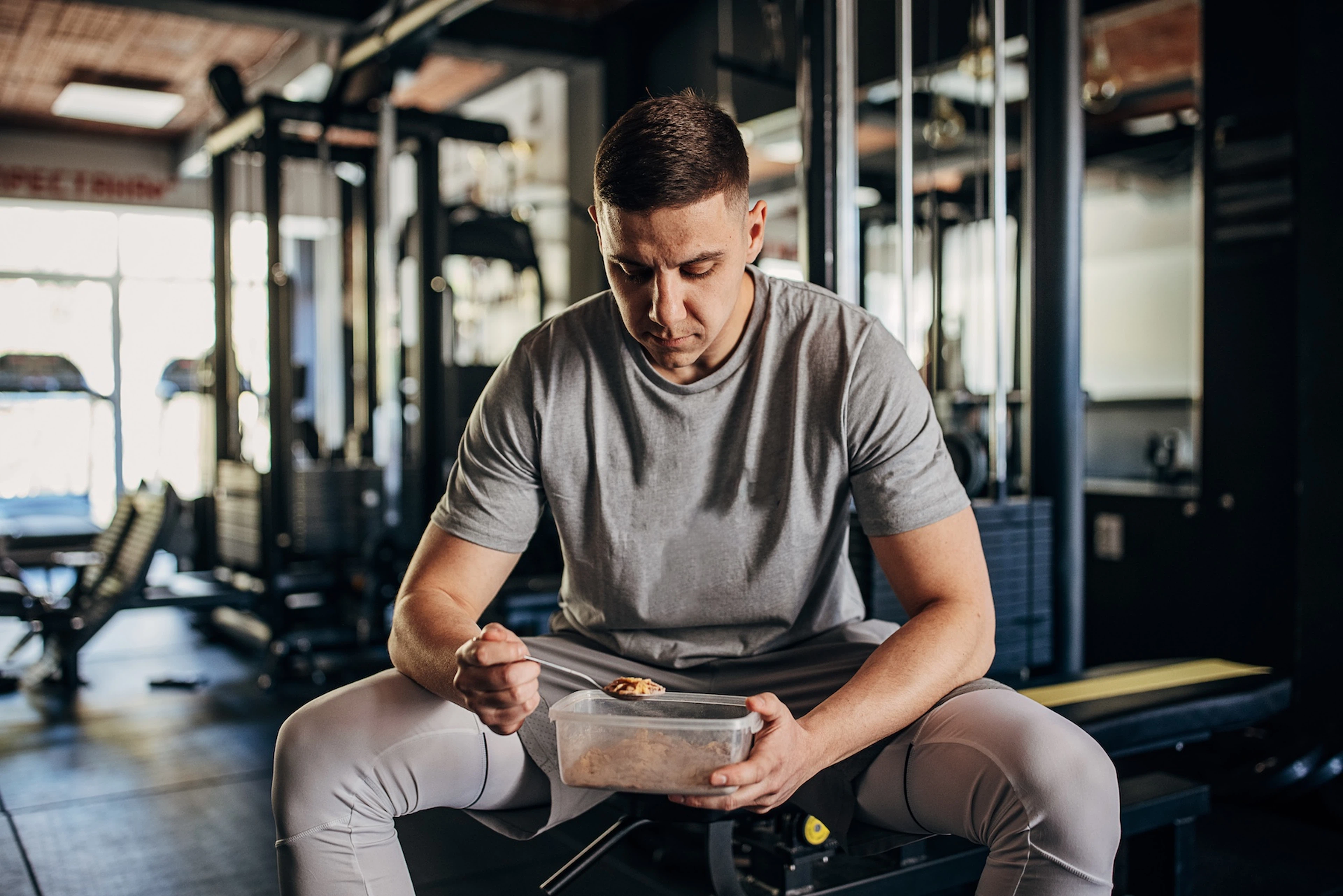 A man eating carbs in order to build muscle. He's sitting on a workout bench eating oats from a plastic container.