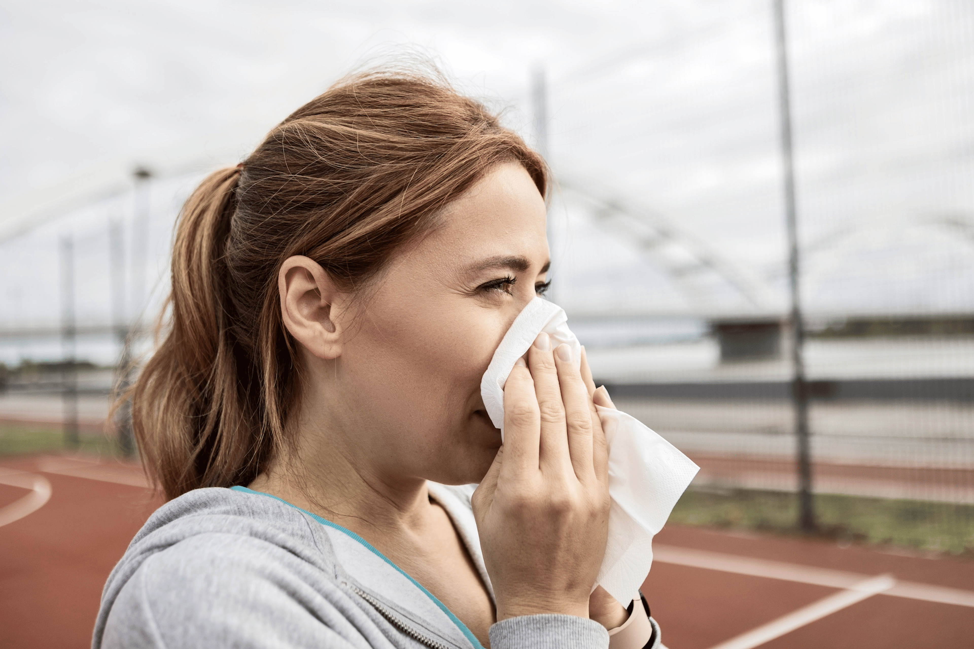 In this image for an article about whether or not you should exercise when you're sick, a woman is blowing her nose into a tissue while she stands outside on a sports court of some kind.