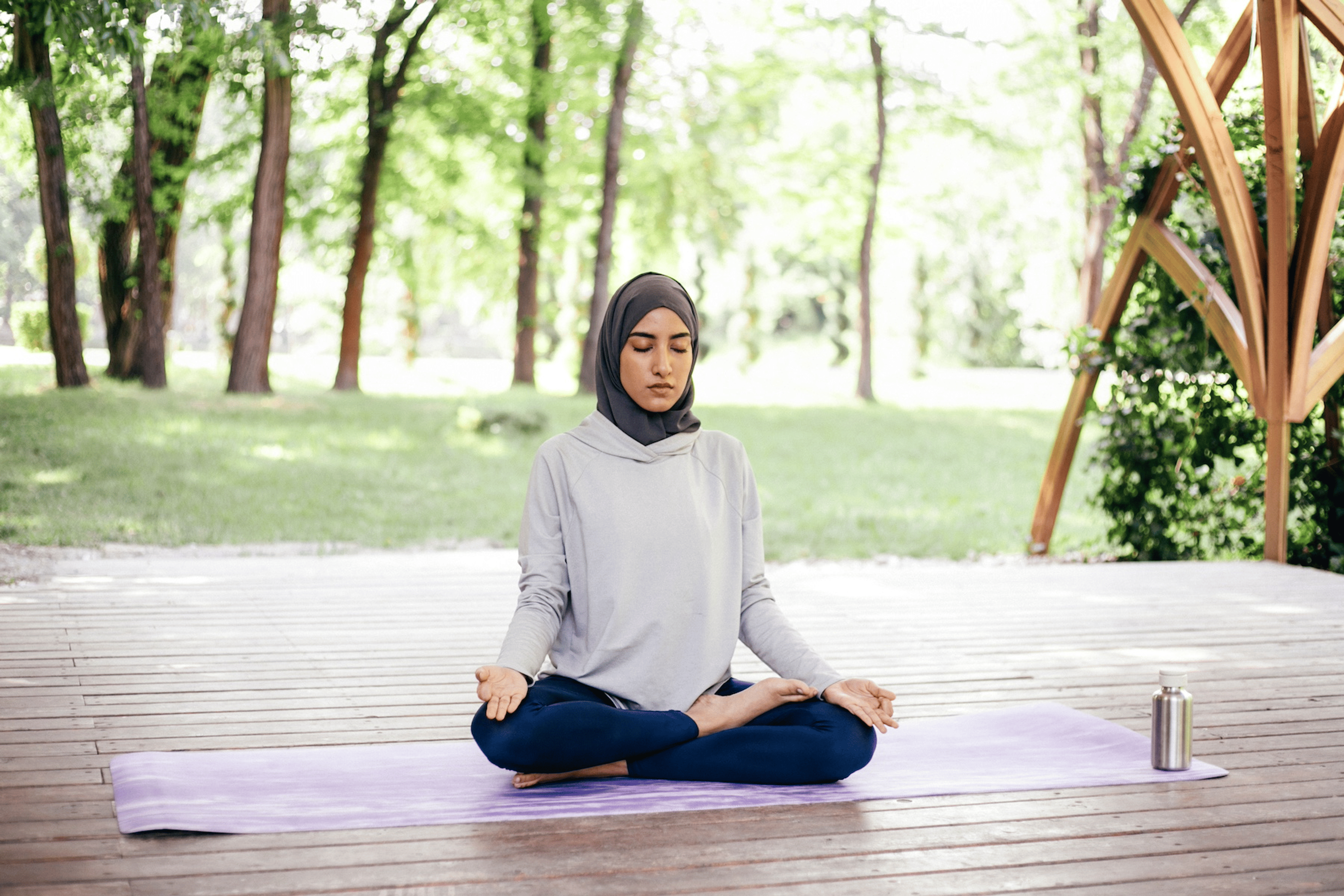 A woman meditating and practicing mindfulness while sitting on a yoga mat in a cross-legged position at home outdoors.