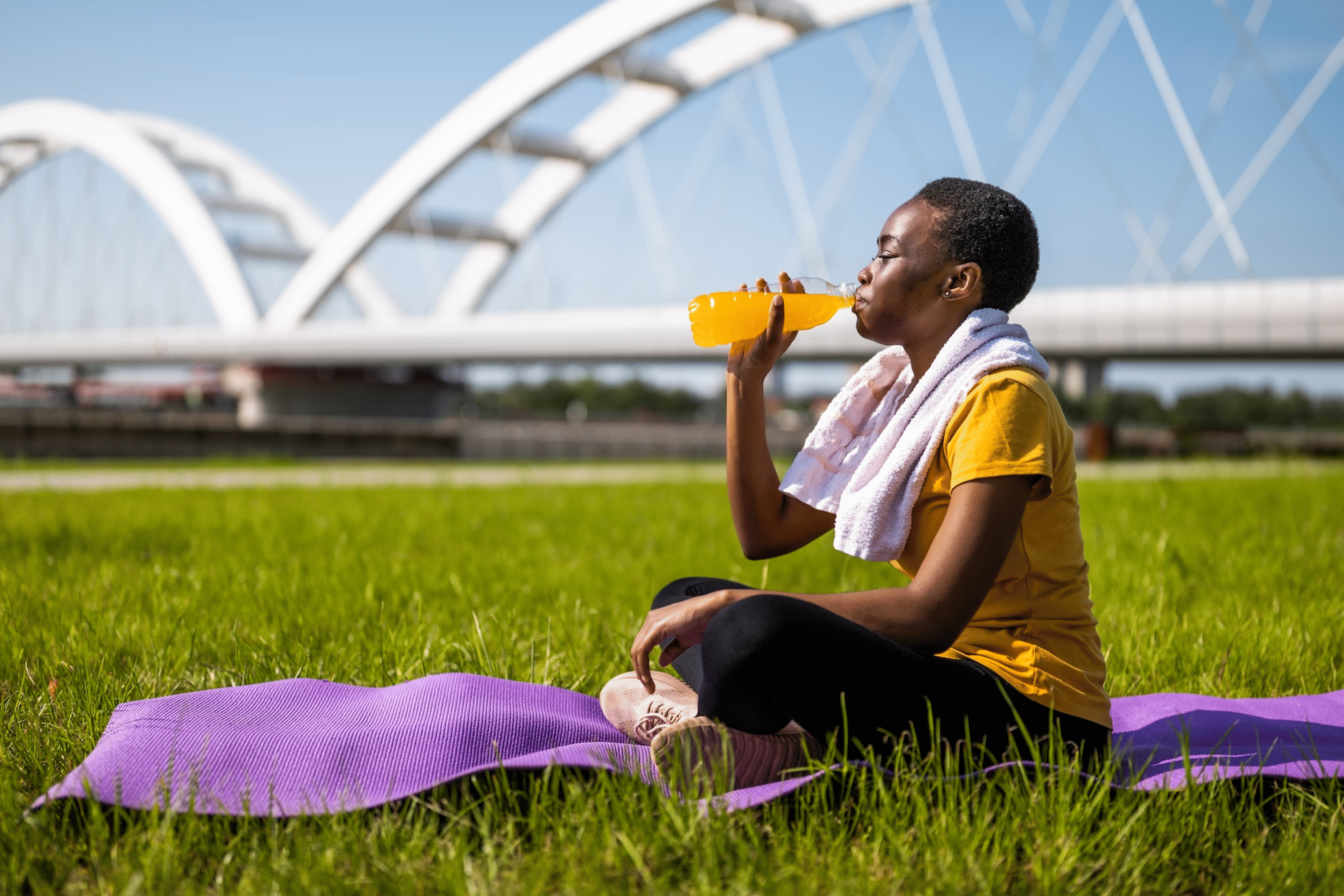 A woman sitting down on a purple yoga mat, closing her eyes and drinking an orange electrolyte drink. She's in an outdoor grassy park and there is a white bridge in the background.